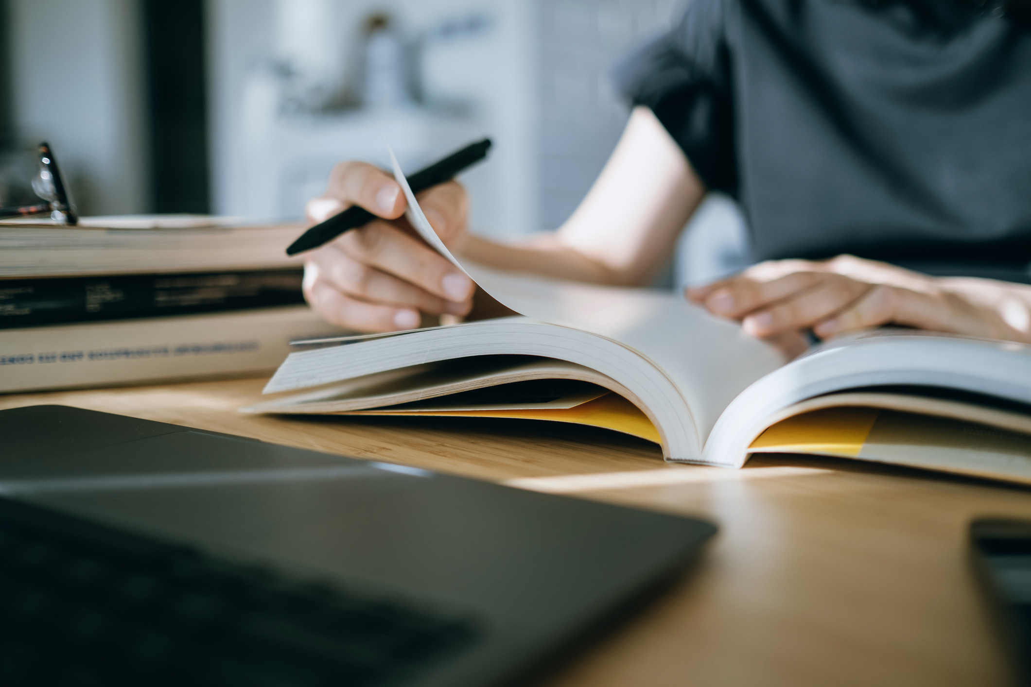 Person in a black shirt writing in an open book at a desk with a laptop and stack of books nearby