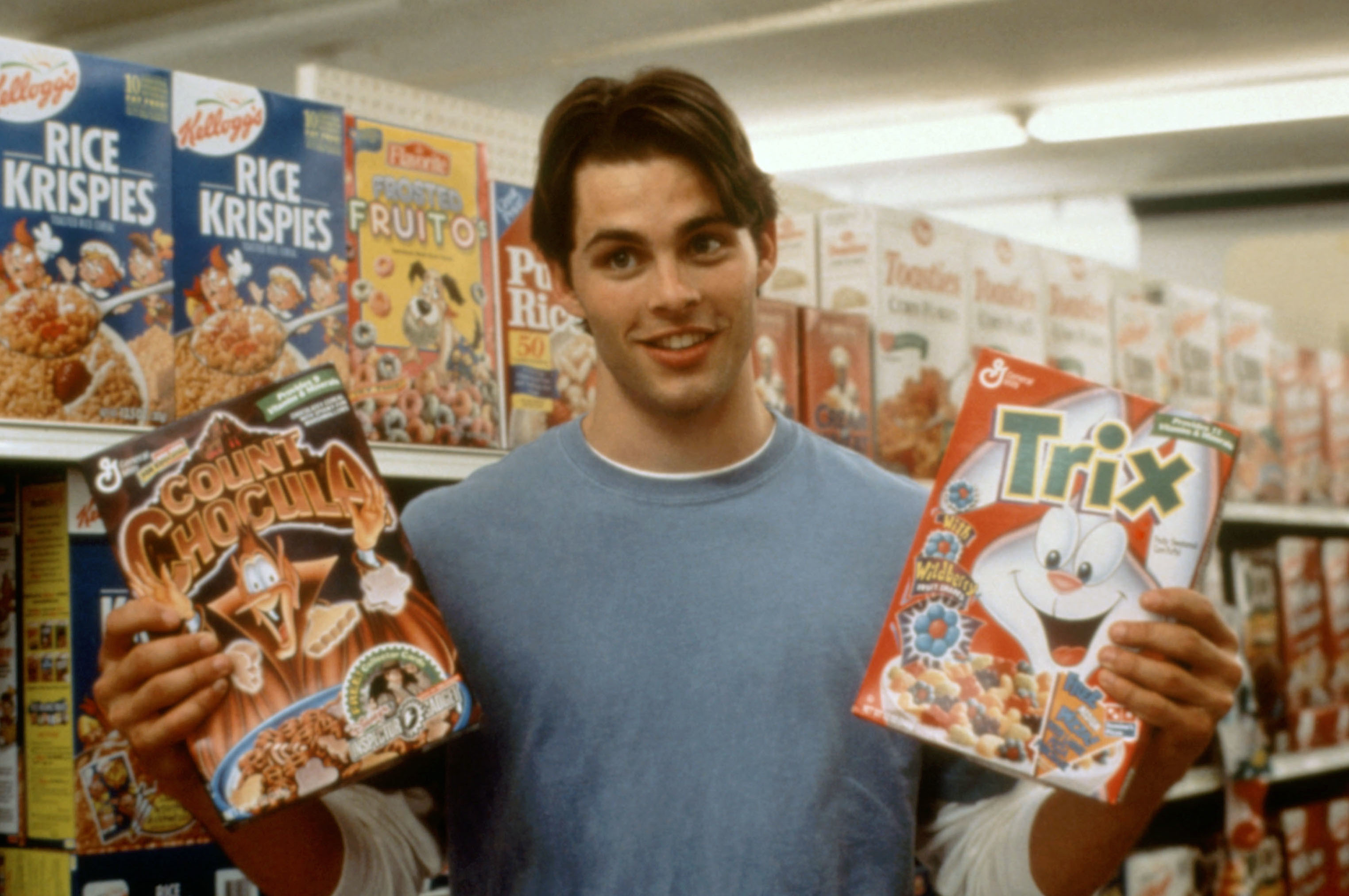 Person in a grocery store aisle holding boxes of Count Chocula and Trix cereal. Shelves filled with various cereals are visible in the background