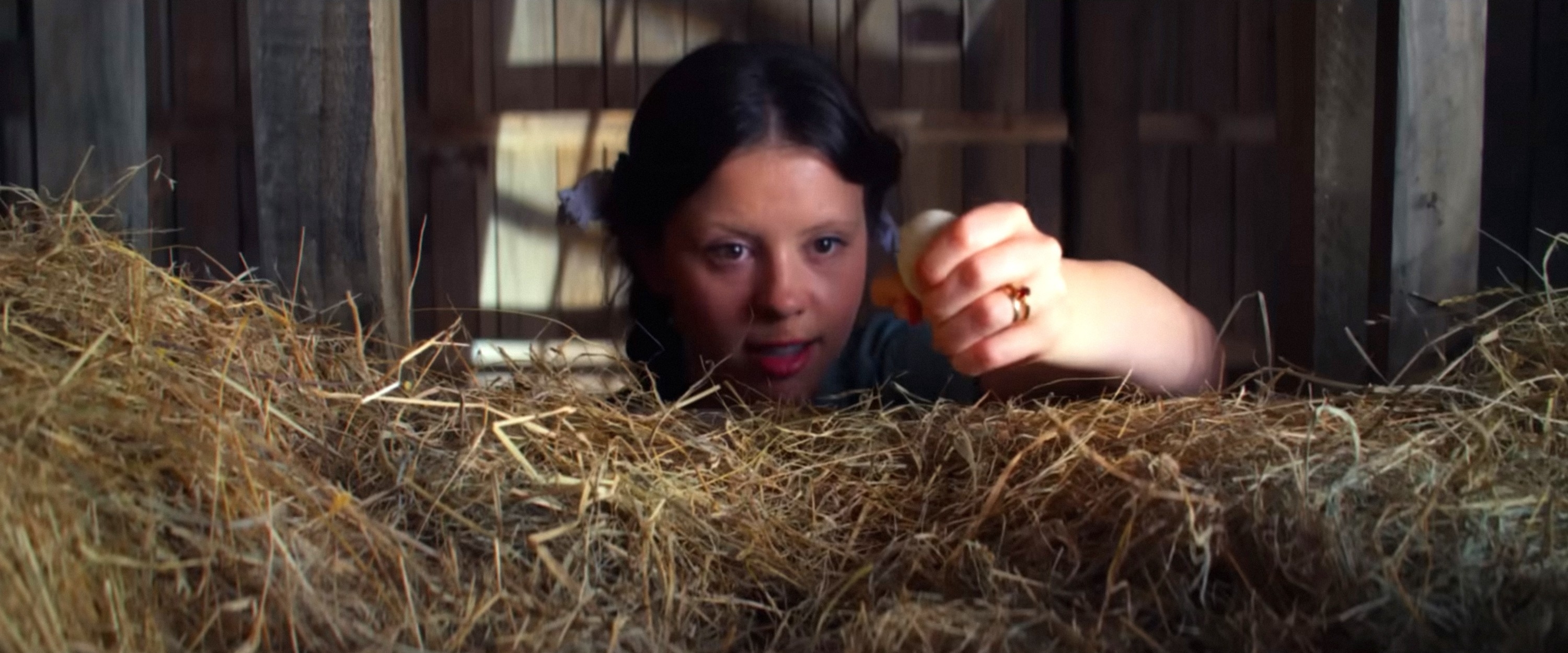 Mia Goth, wearing a simple dress, reaches out towards an object in a barn filled with hay, with a determined expression on her face