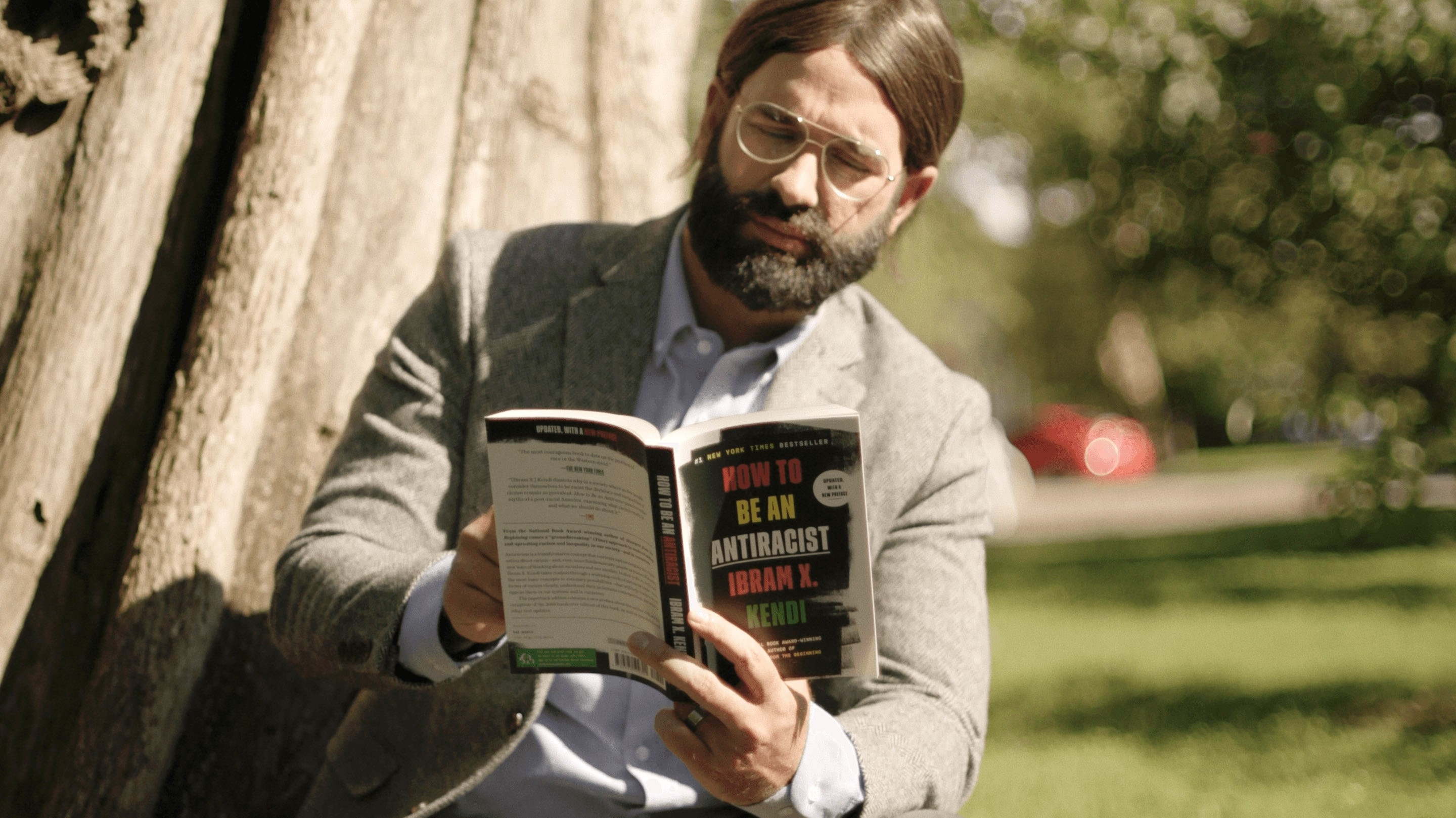 A person with glasses and a beard is sitting by a tree, reading the book "How to Be an Antiracist" by Ibram X. Kendi