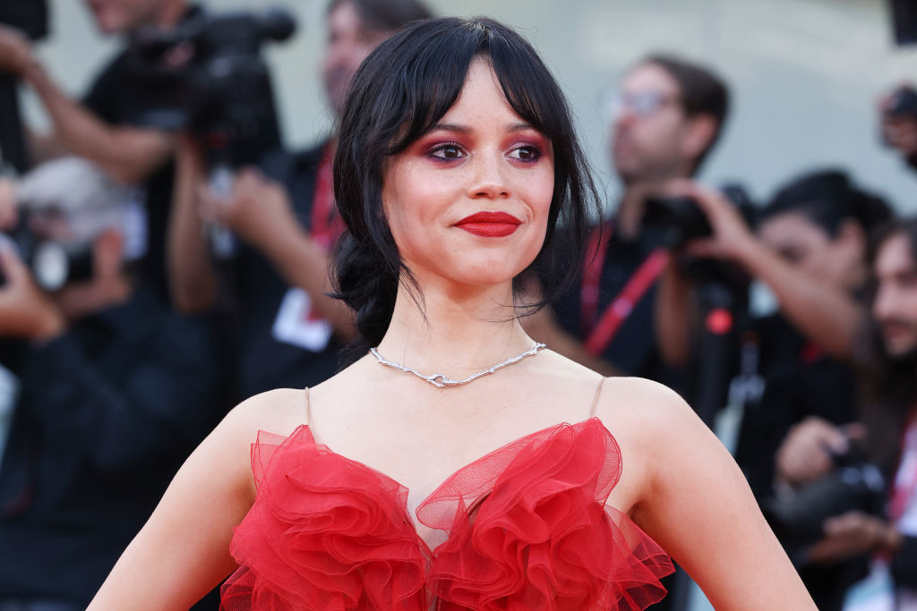 Jenna Ortega poses on the red carpet wearing a sheer, ruffled red dress, with photographers in the background