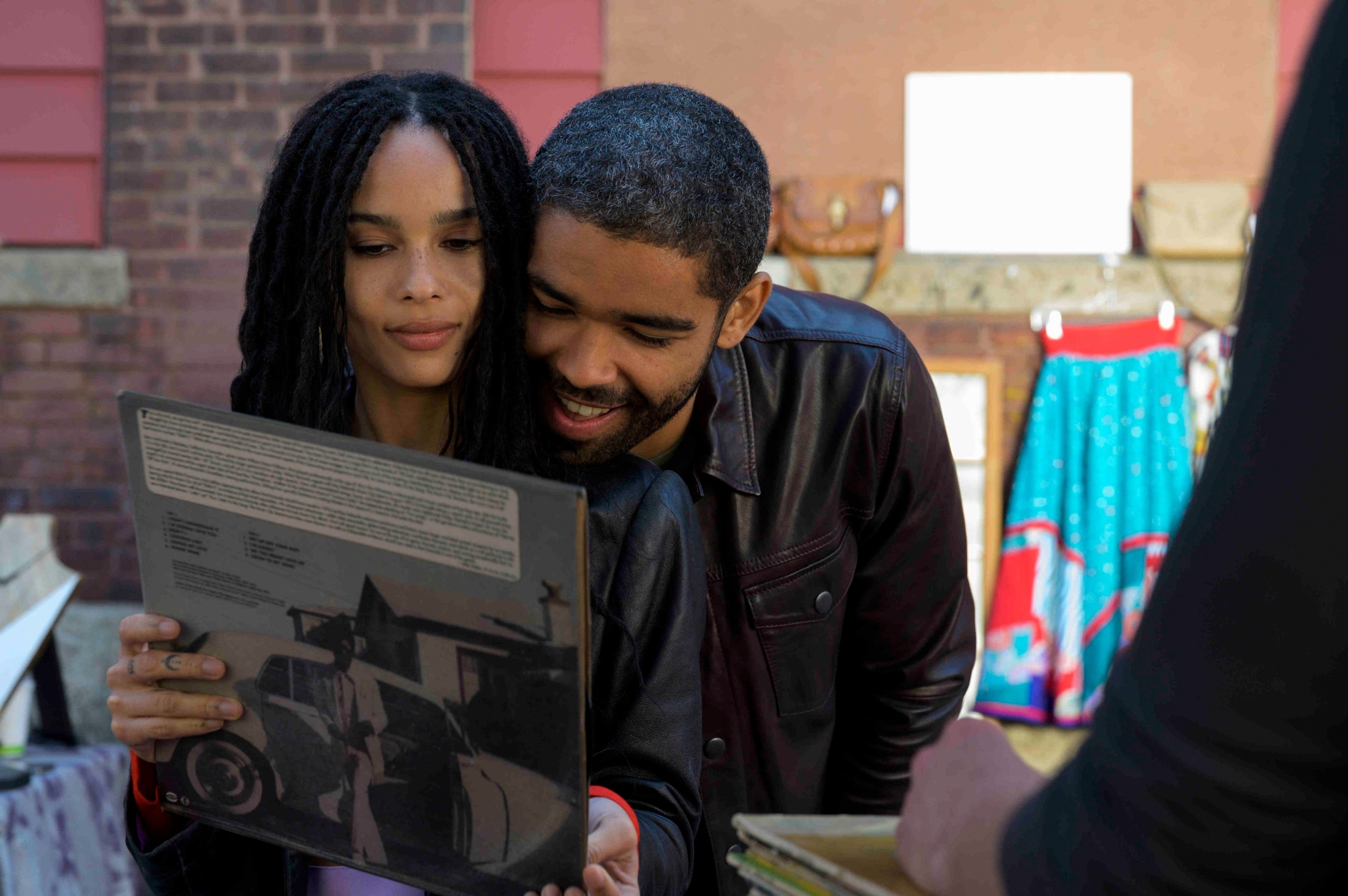 Zoë Kravitz and co-star reading the back of a vinyl record together, dressed casually, on a street with market stalls in the background