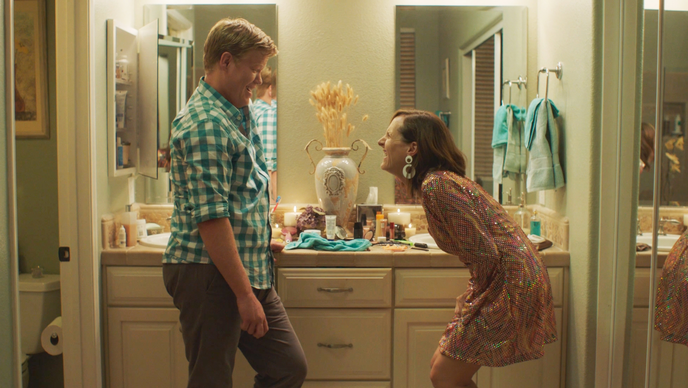 Kristen Wiig in a colorful, patterned dress leaning forward and laughing with a man in a blue plaid shirt in a bathroom