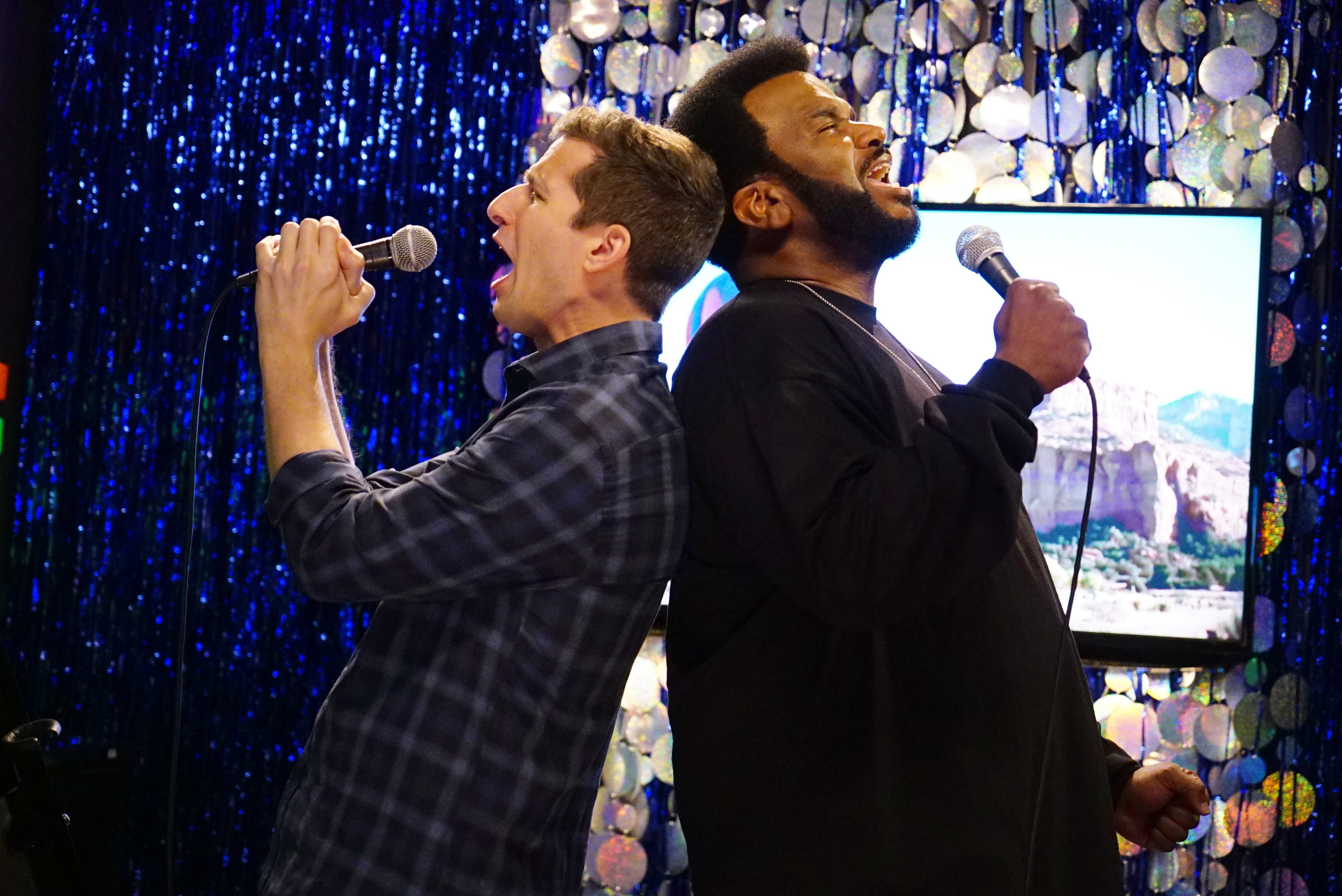 Andy Samberg and Craig Robinson are standing back-to-back, singing into microphones on a stage with a shimmering, decorative backdrop