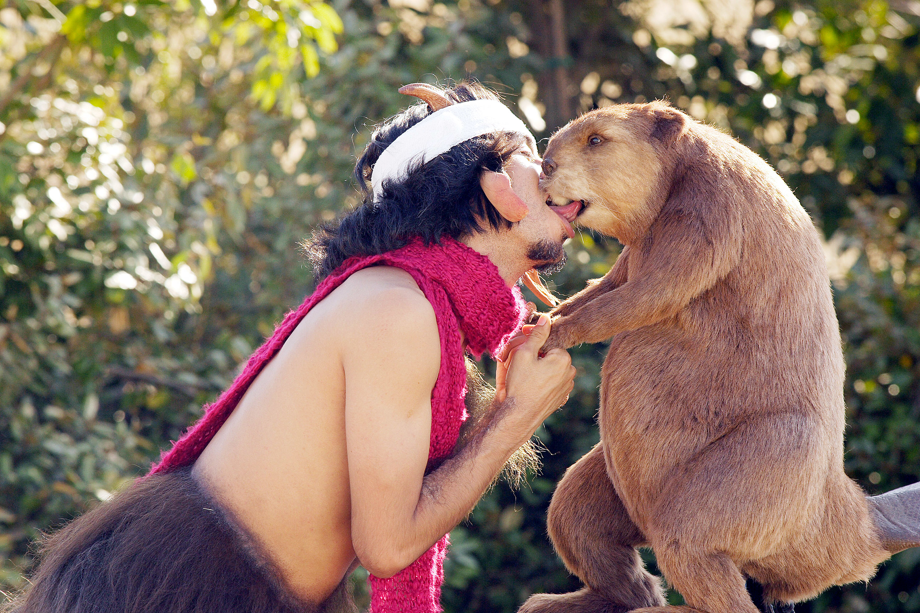 A person dressed as a satyr, wearing a headband and pink scarf, receives a kiss from a large animal, likely a mammal, in an outdoor setting