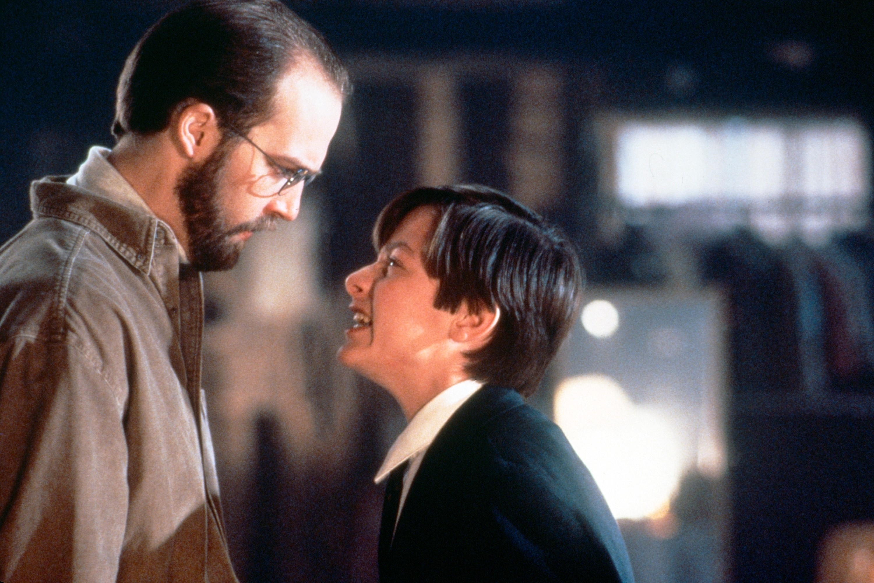 A man in glasses and a brown jacket stares down at a person in a dark blazer and white shirt who is angrily yelling at him