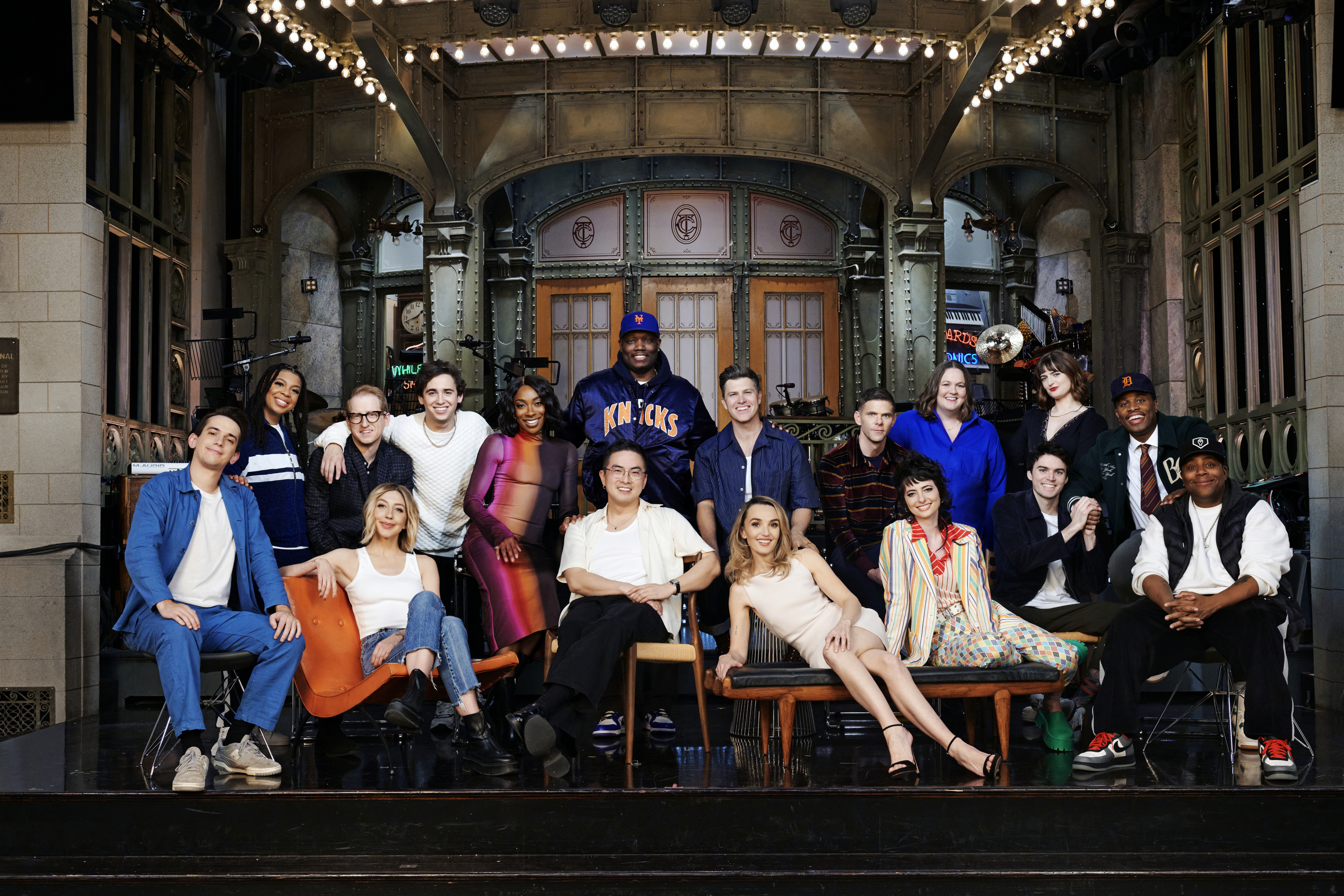 The image shows the cast of Saturday Night Live posed together on stage in casual and semi-formal clothing