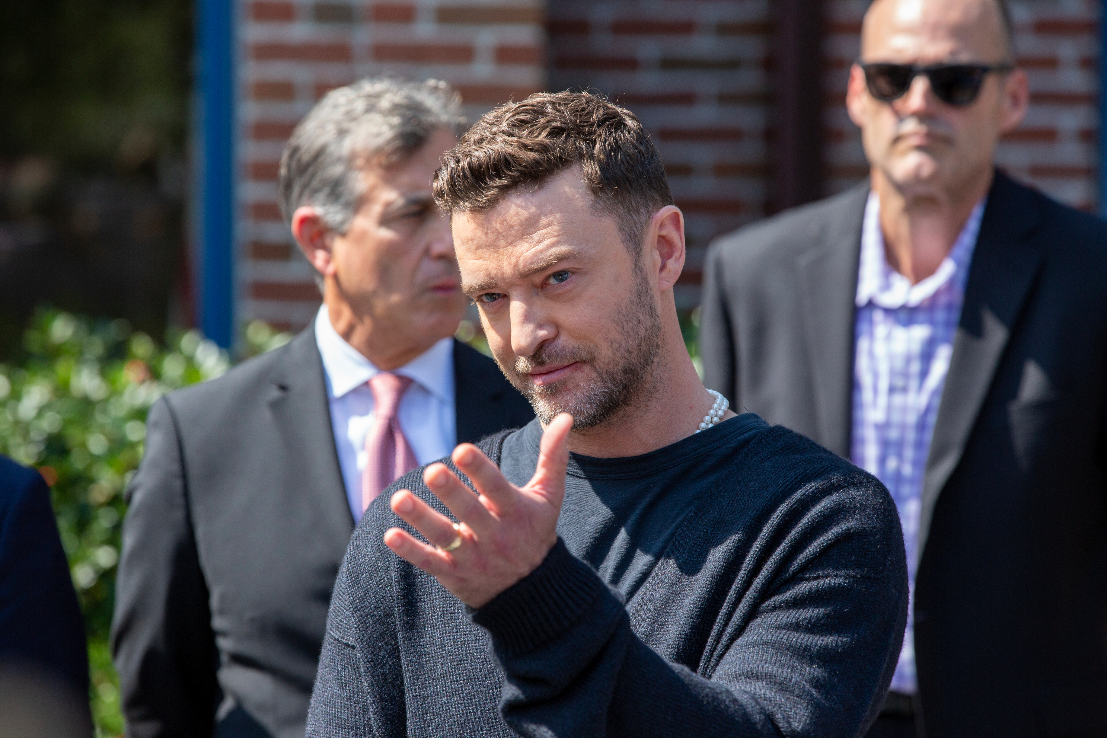 Justin Timberlake stands in front of two suited men, gesturing with his hand at an event outdoors