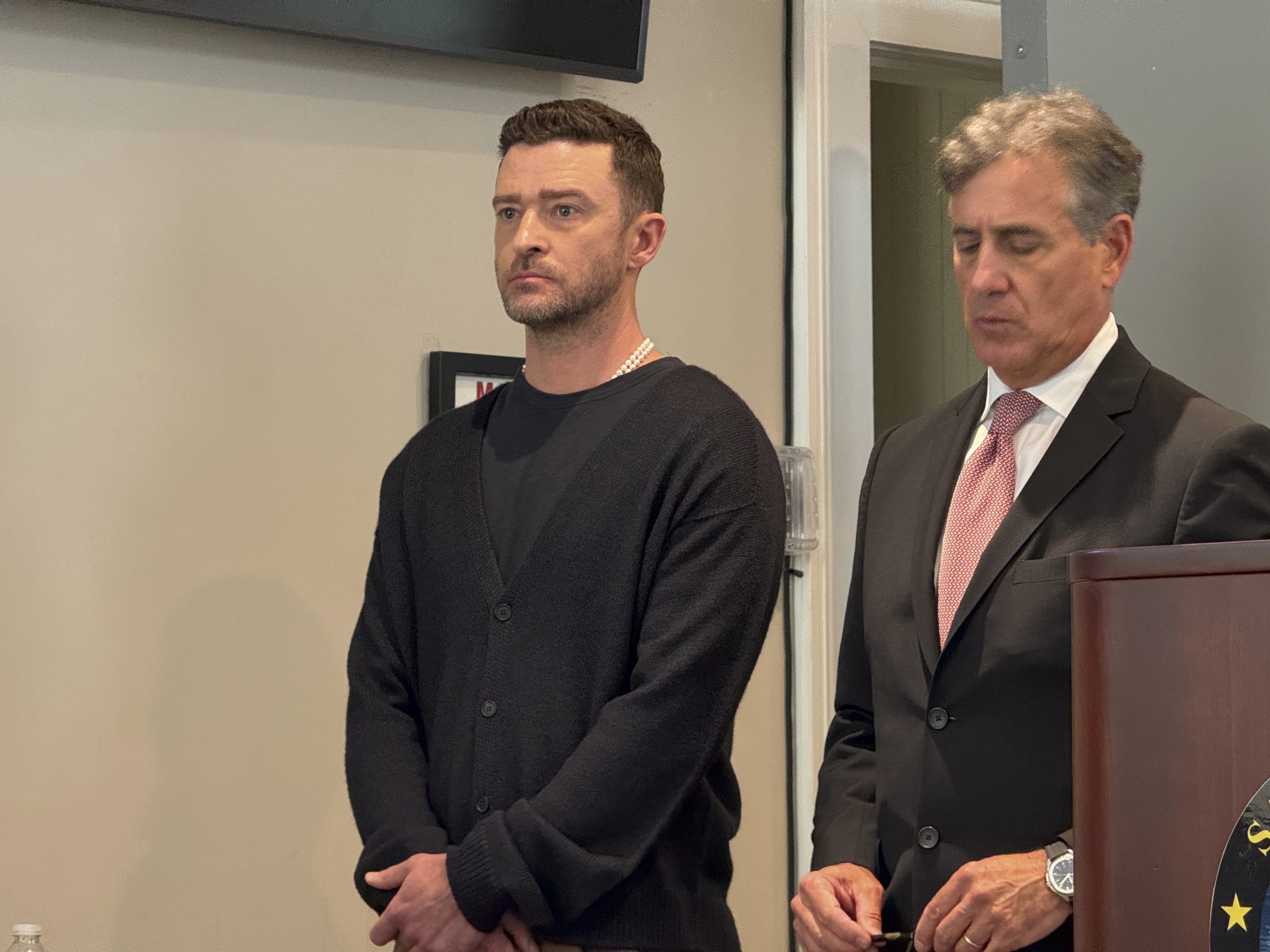 Justin Timberlake stands next to a man in a suit at a formal event. Timberlake wears a casual sweater and shirt