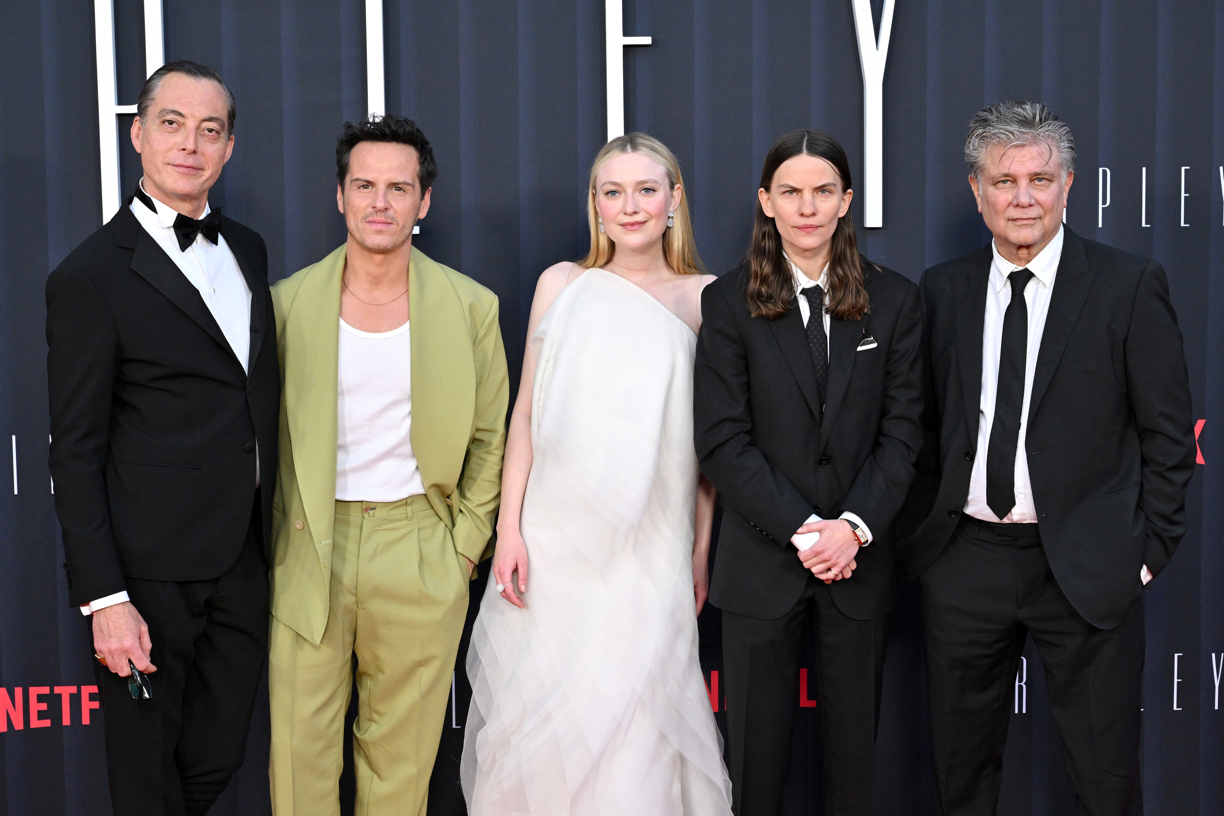 Five individuals pose on the red carpet at a Netflix event. Men in tuxedos; Dakota in an elegant one-shoulder gown
