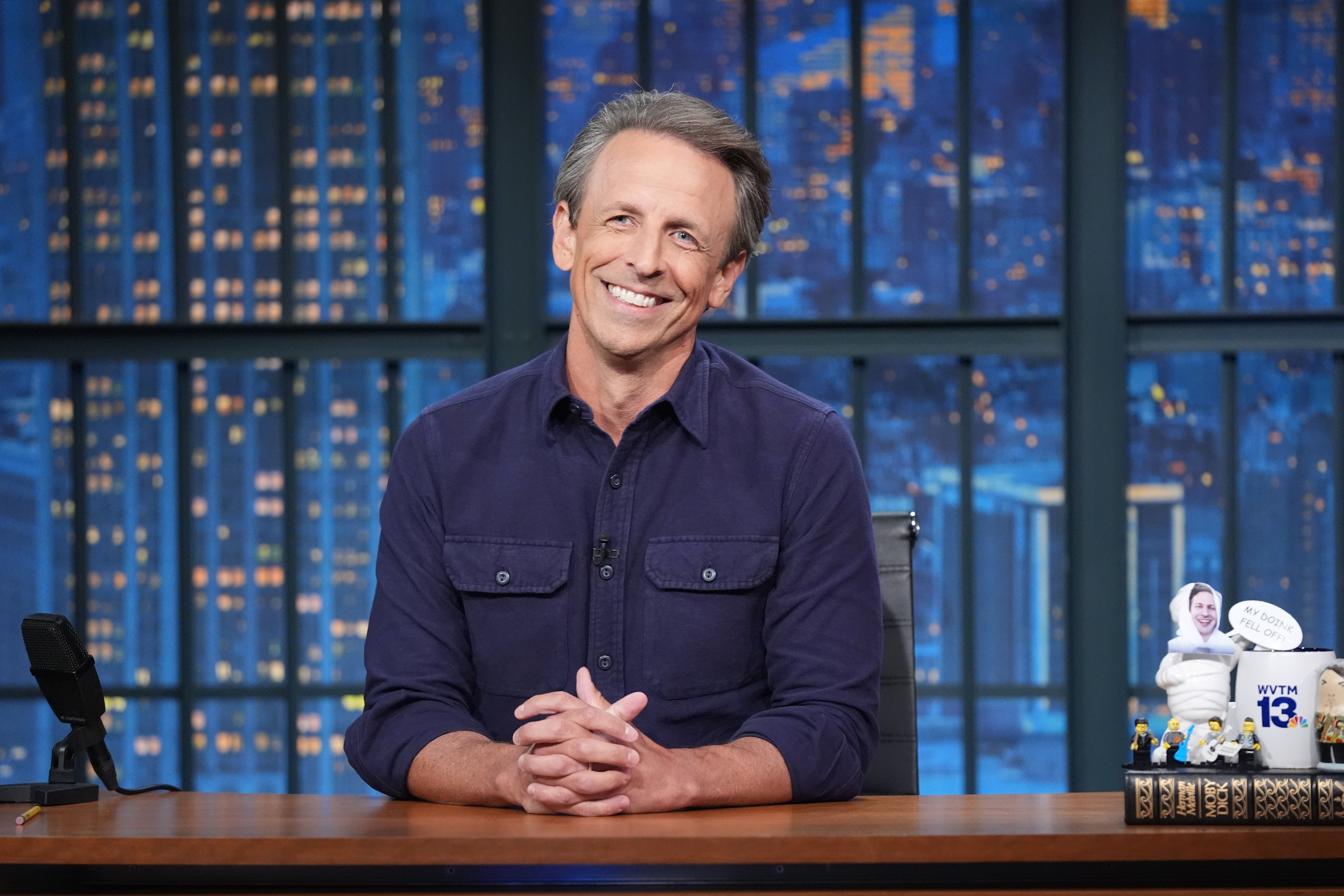 Seth Meyers sits at his desk on the set of "Late Night with Seth Meyers," smiling and wearing a casual button-down shirt with New York City buildings in the background