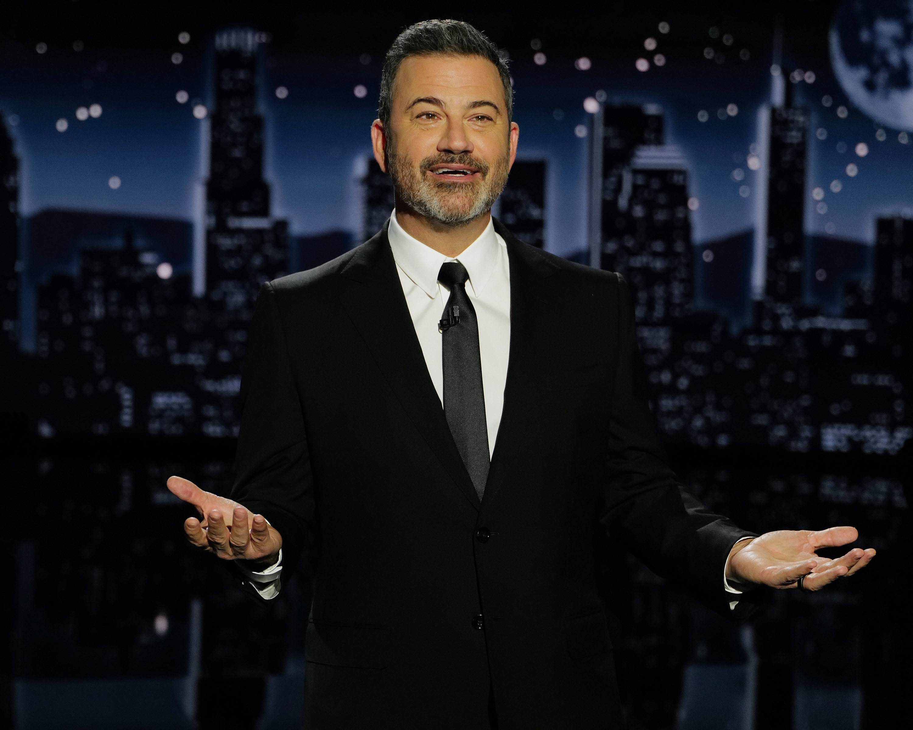 Jimmy Kimmel, wearing a suit and tie, gestures while speaking on stage with a city skyline backdrop at night