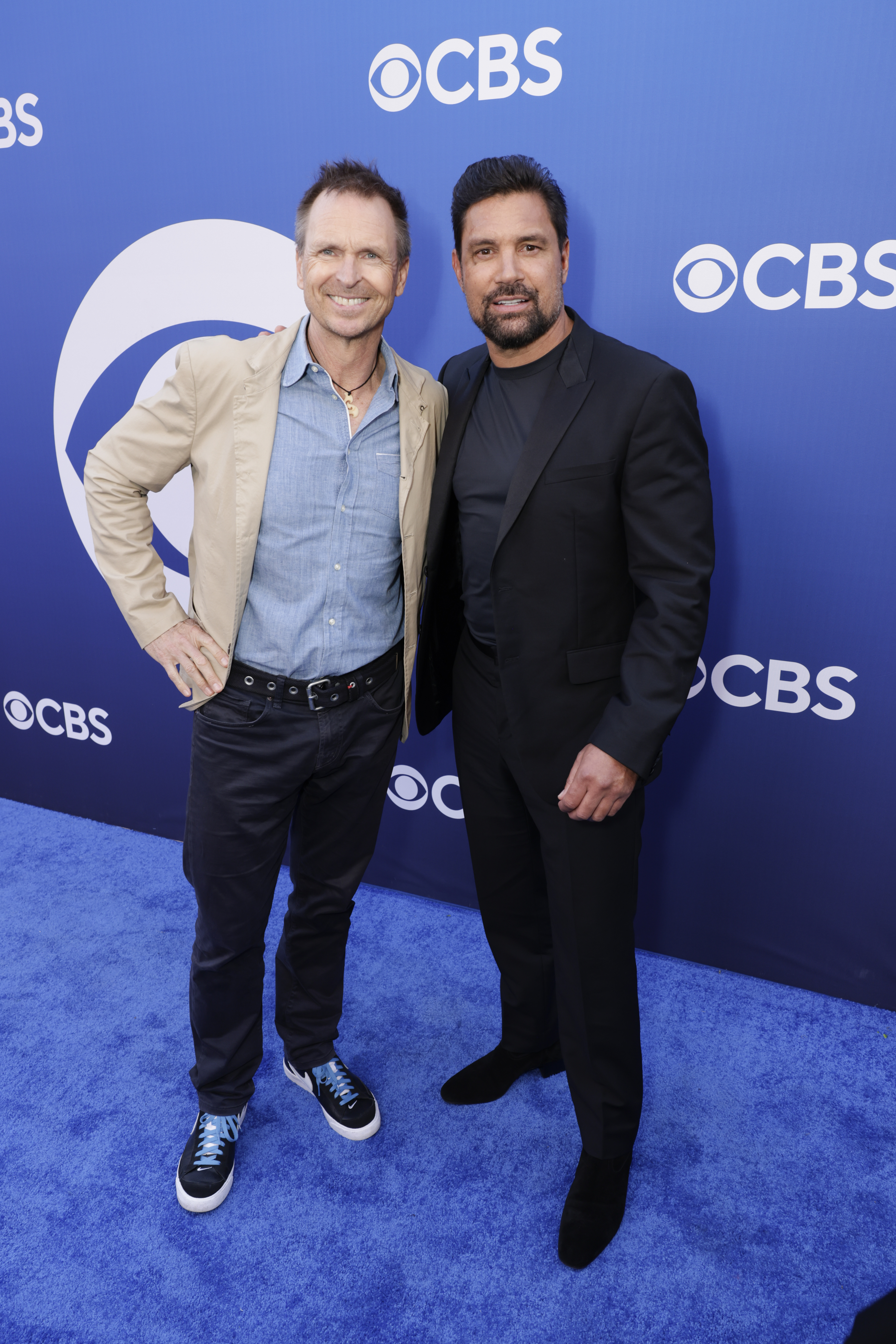 Two men standing on a blue carpet, smiling at a CBS event. One wears a tan jacket over a blue shirt and jeans, and the other wears a black suit.