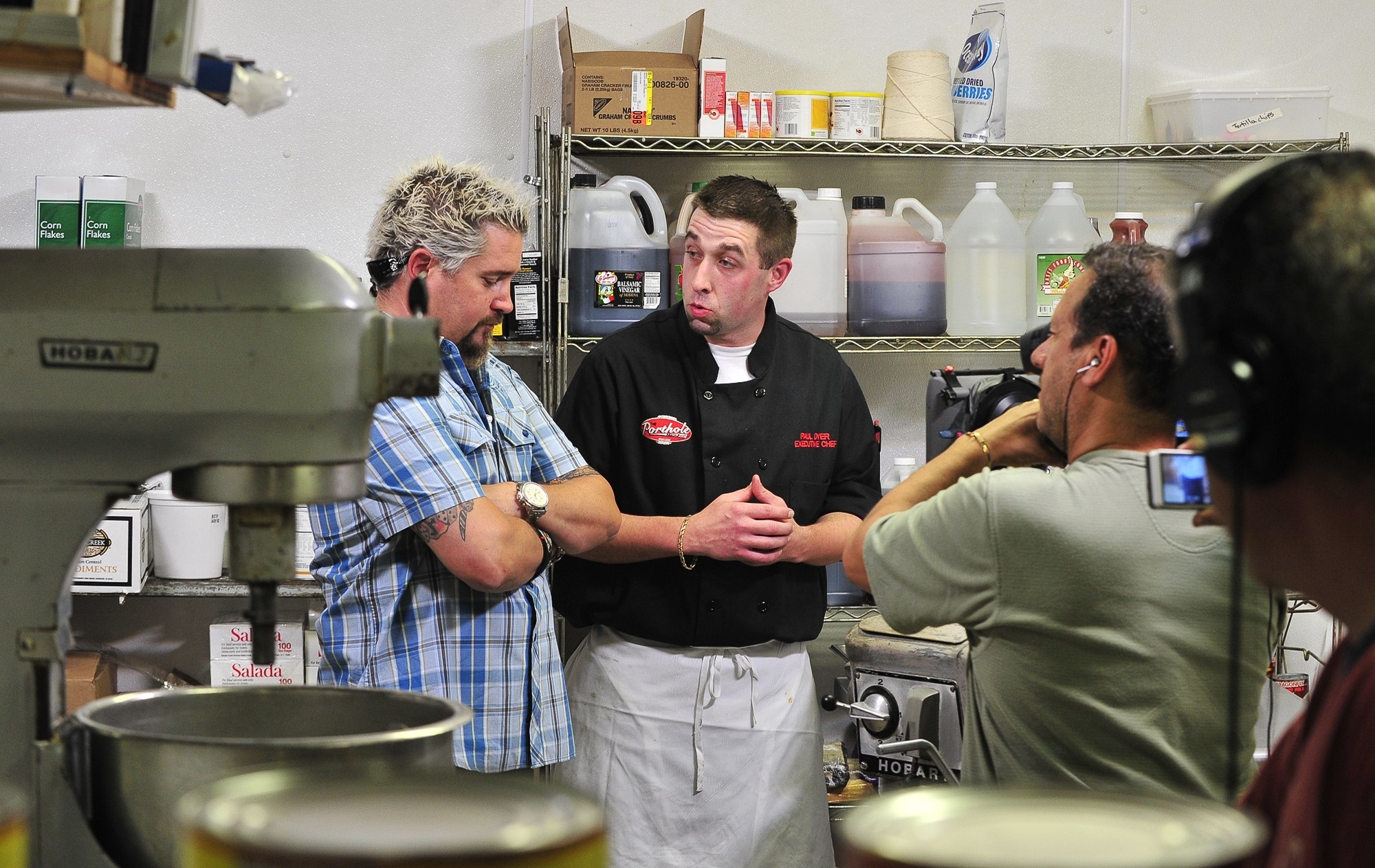 Guy Fieri, dressed casually, talks to two men in a professional kitchen. One man is wearing a chef's jacket and apron, and the other holds a video camera
