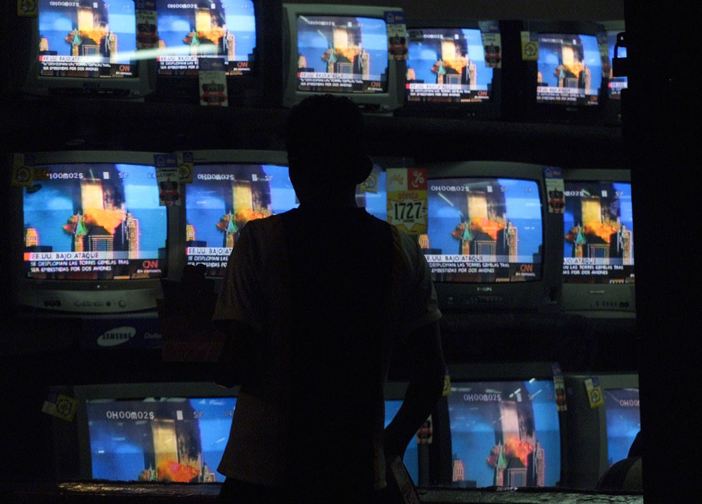 A person stands in front of multiple TV screens, all displaying news footage of the World Trade Center attack on 9/11
