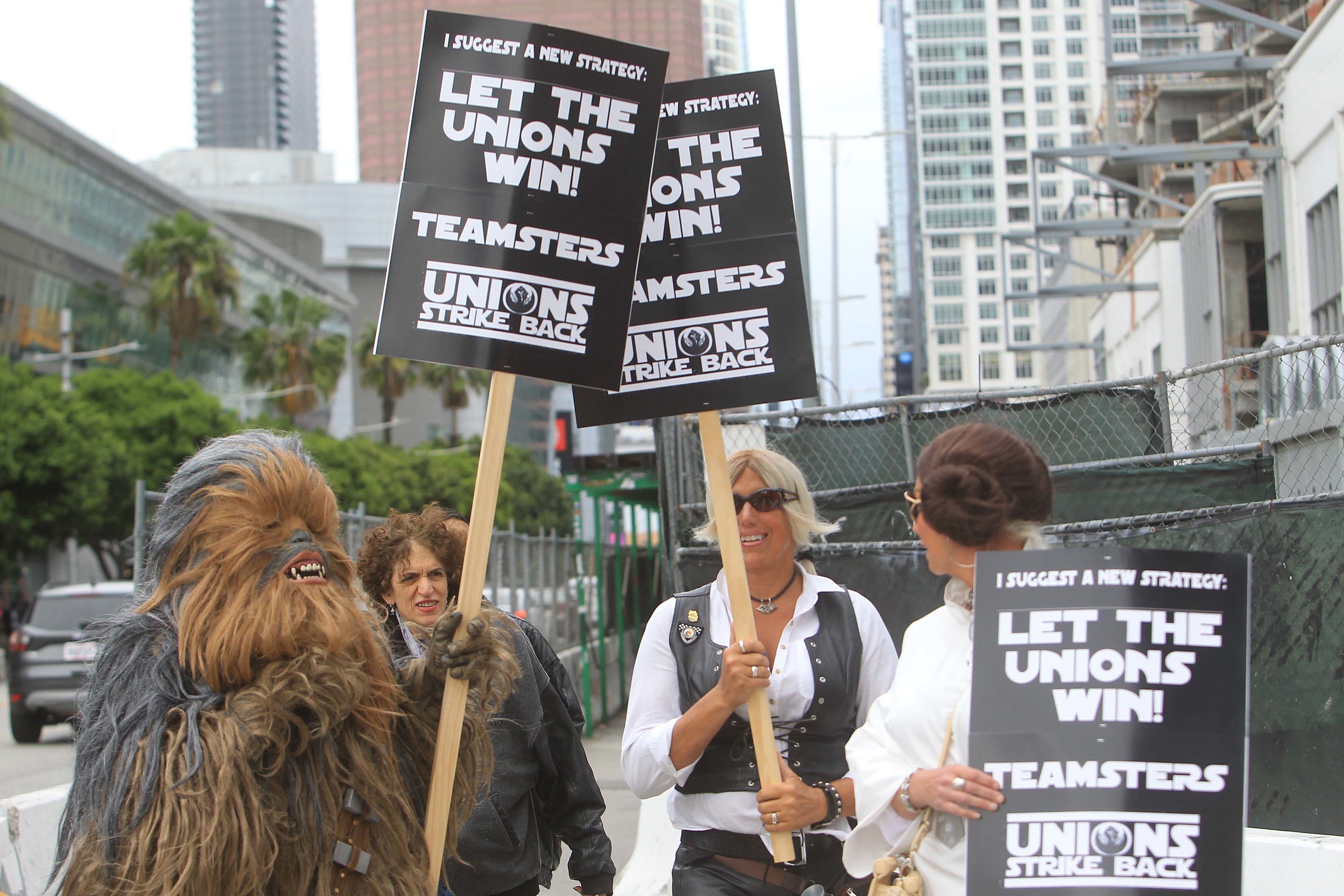 Chewbacca, along with other individuals holding signs that say "LET THE UNIONS WIN!" and "UNIONS STRIKE BACK" in a city street protest