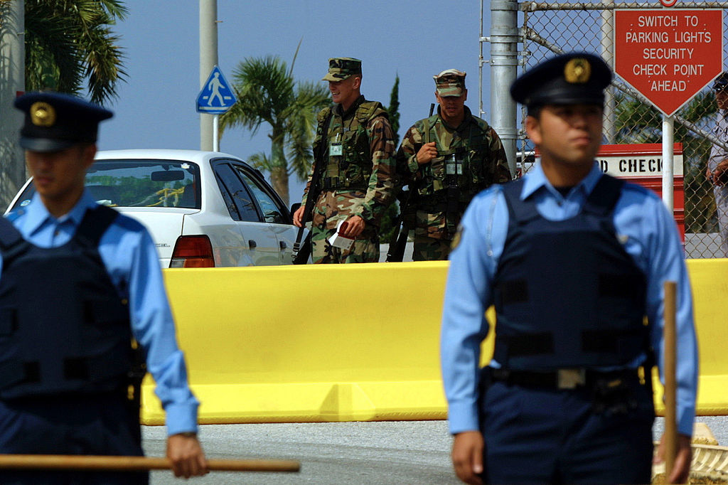 Military personnel in camouflage and armed security officers stand guard at a checkpoint with a yellow barricade and a sign about parking lights and security ahead
