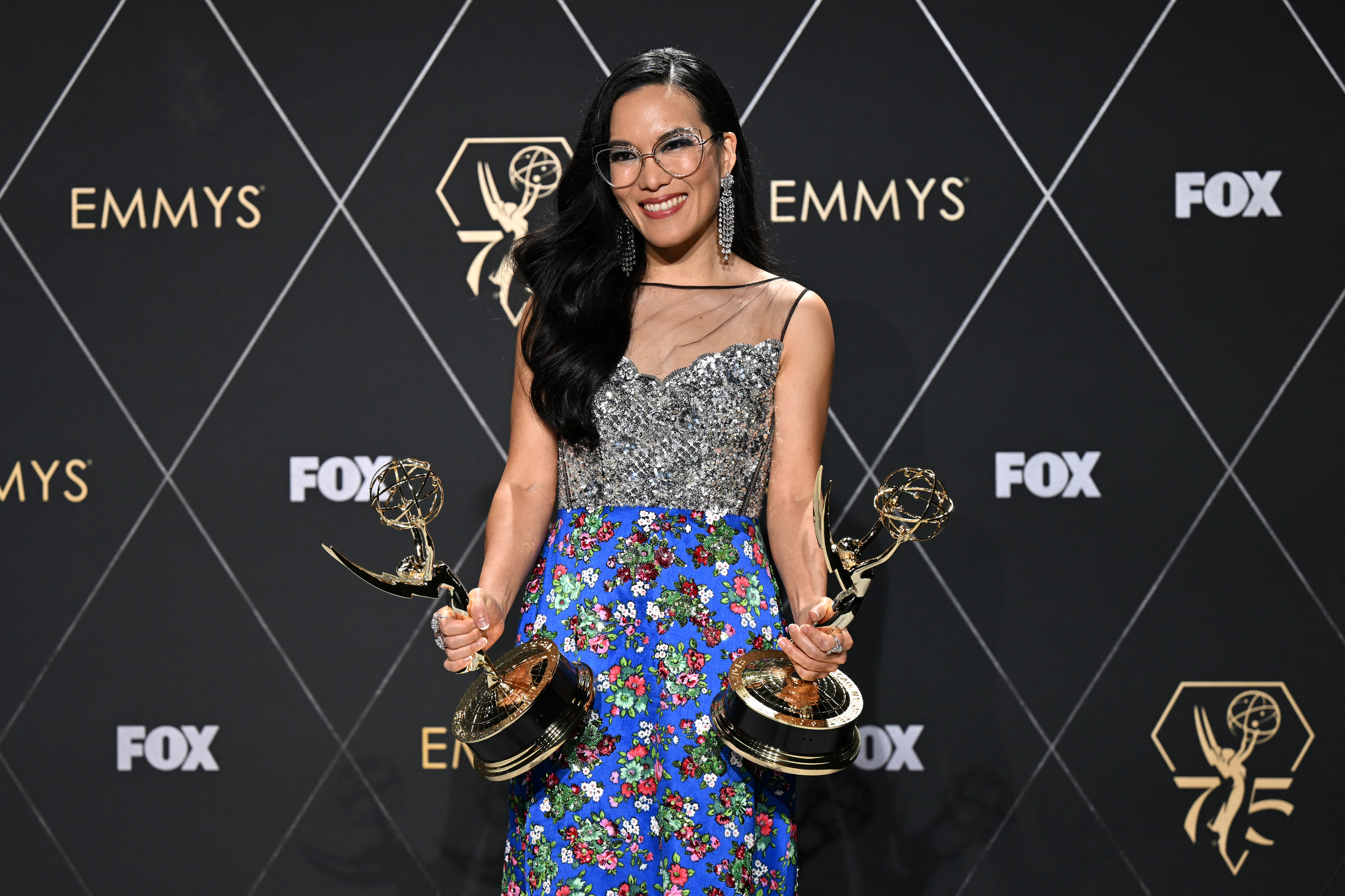 Ali Wong smiles while holding two Emmy awards. She is wearing a sparkling top and a floral skirt