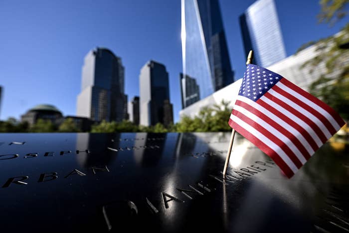 An American flag is planted on a memorial with blurred city buildings in the background, symbolizing remembrance. Names are inscribed on the memorial