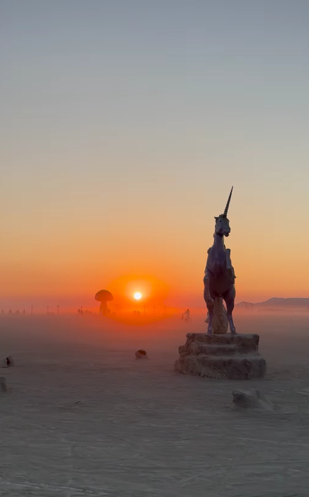 A unicorn statue stands in a desert landscape at sunrise at Burning Man, with a glowing sun near the horizon and a mushroom cloud-shaped structure in the distance