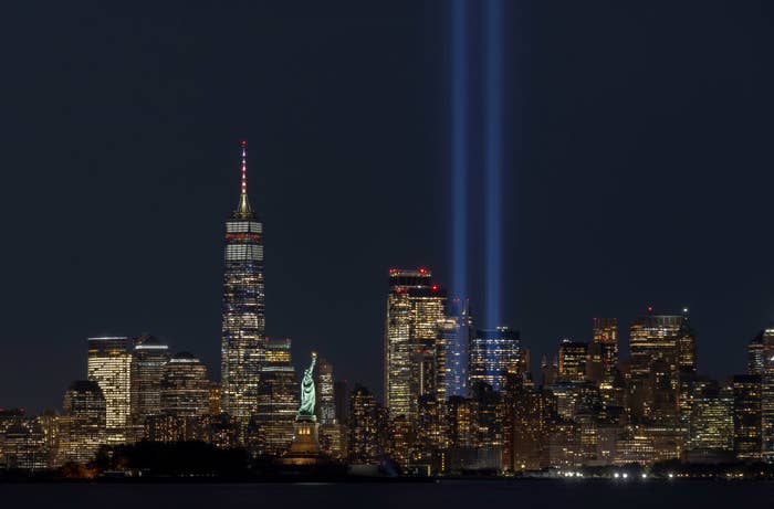 New York City skyline at night, featuring the Tribute in Light memorial with twin beams of light and the Statue of Liberty visible in the foreground