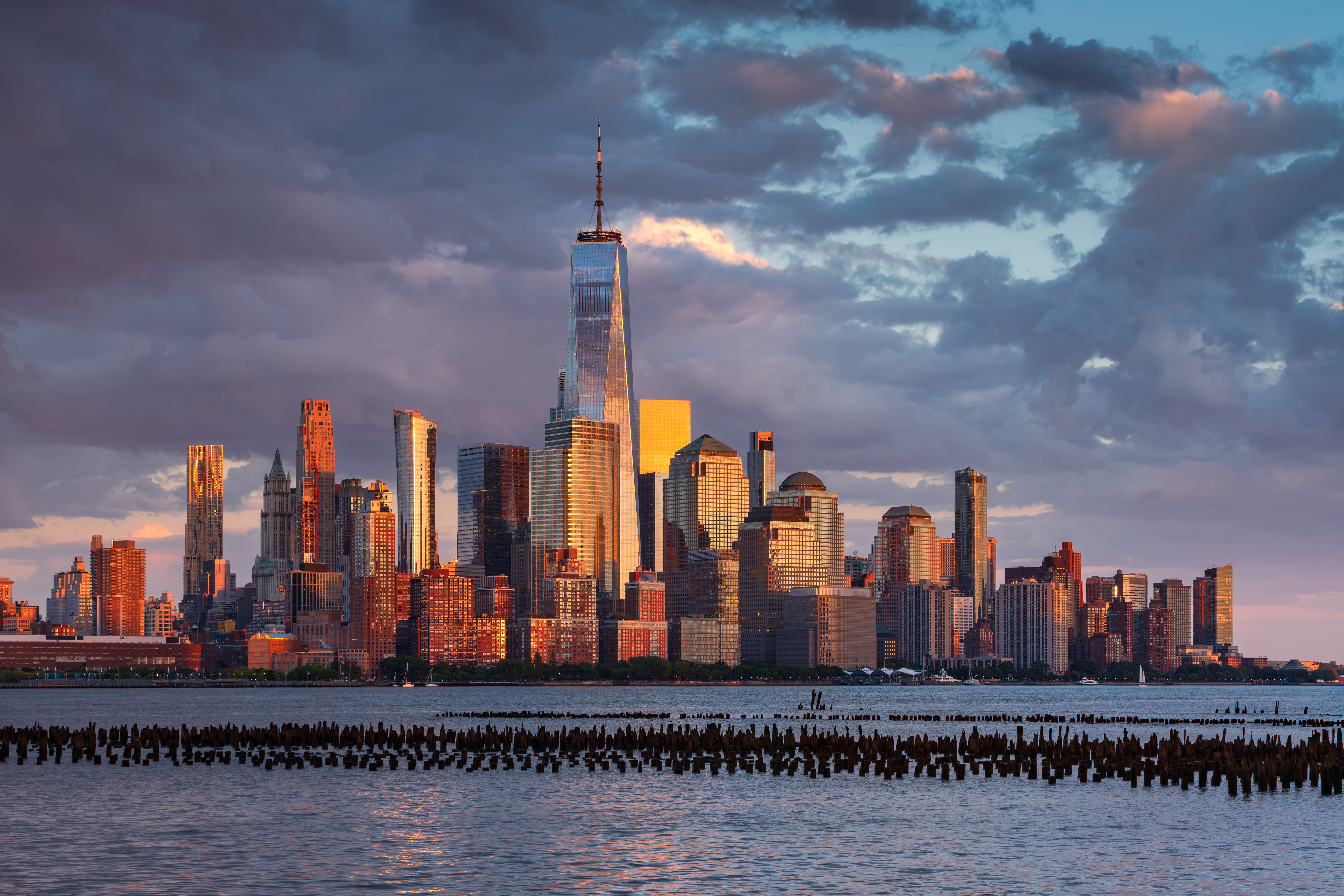 New York City skyline featuring the One World Trade Center and various surrounding buildings as seen from across the waterfront at sunset
