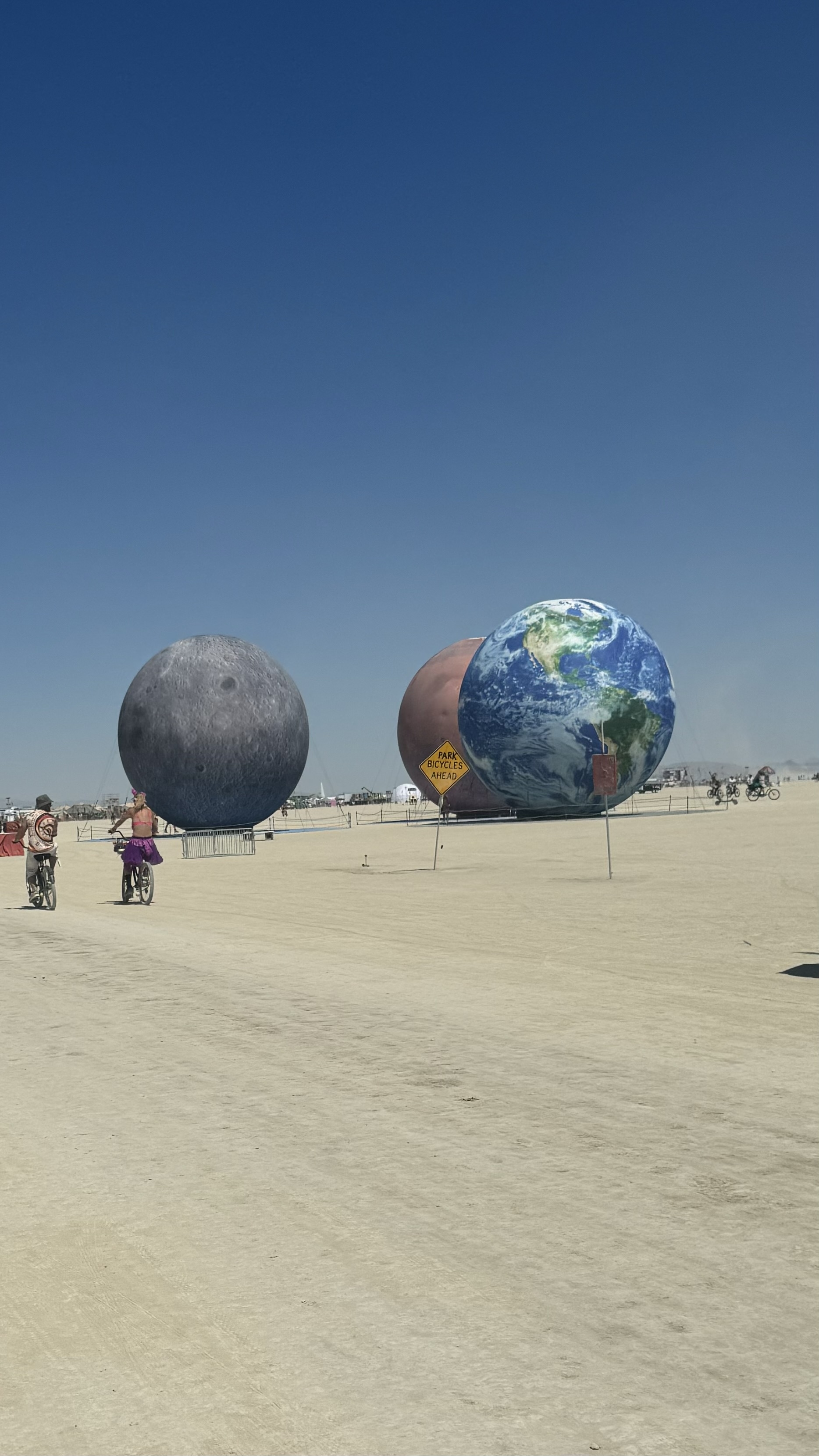 Large model planets at Burning Man including Earth are displayed on a desert expanse under a clear blue sky, with people nearby on bicycles