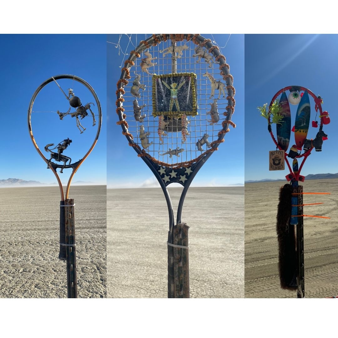 Three decorated tennis racquets displayed outdoors in a desert. Each racquet has unique artistic elements including sculptures, netting, and various other materials