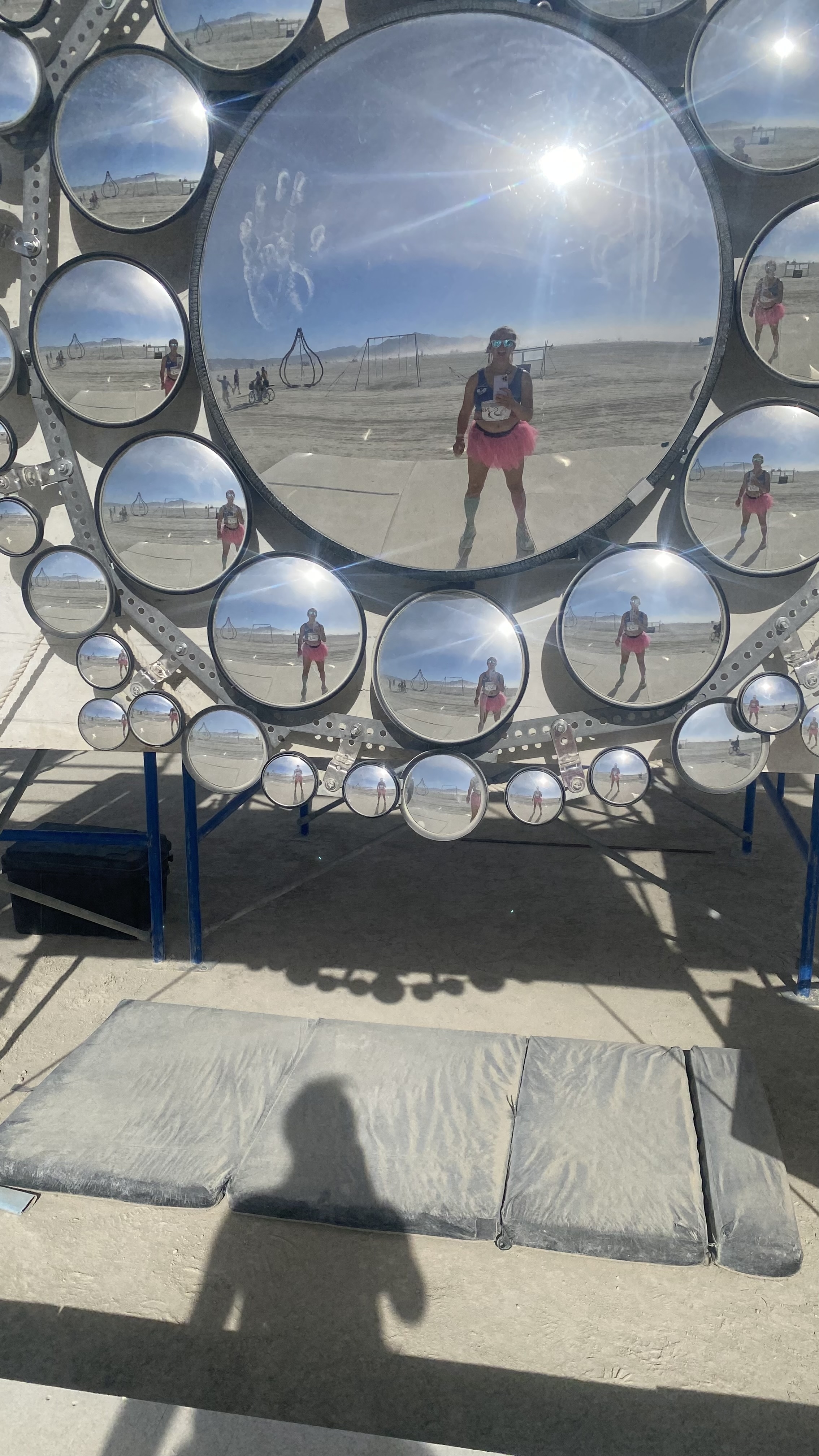 Elizabeth Swaney wearing a tutu and bib during the Burning Man Ultramarathon, reflected in multiple circular mirrors positioned on a metal frame at an outdoor art installation
