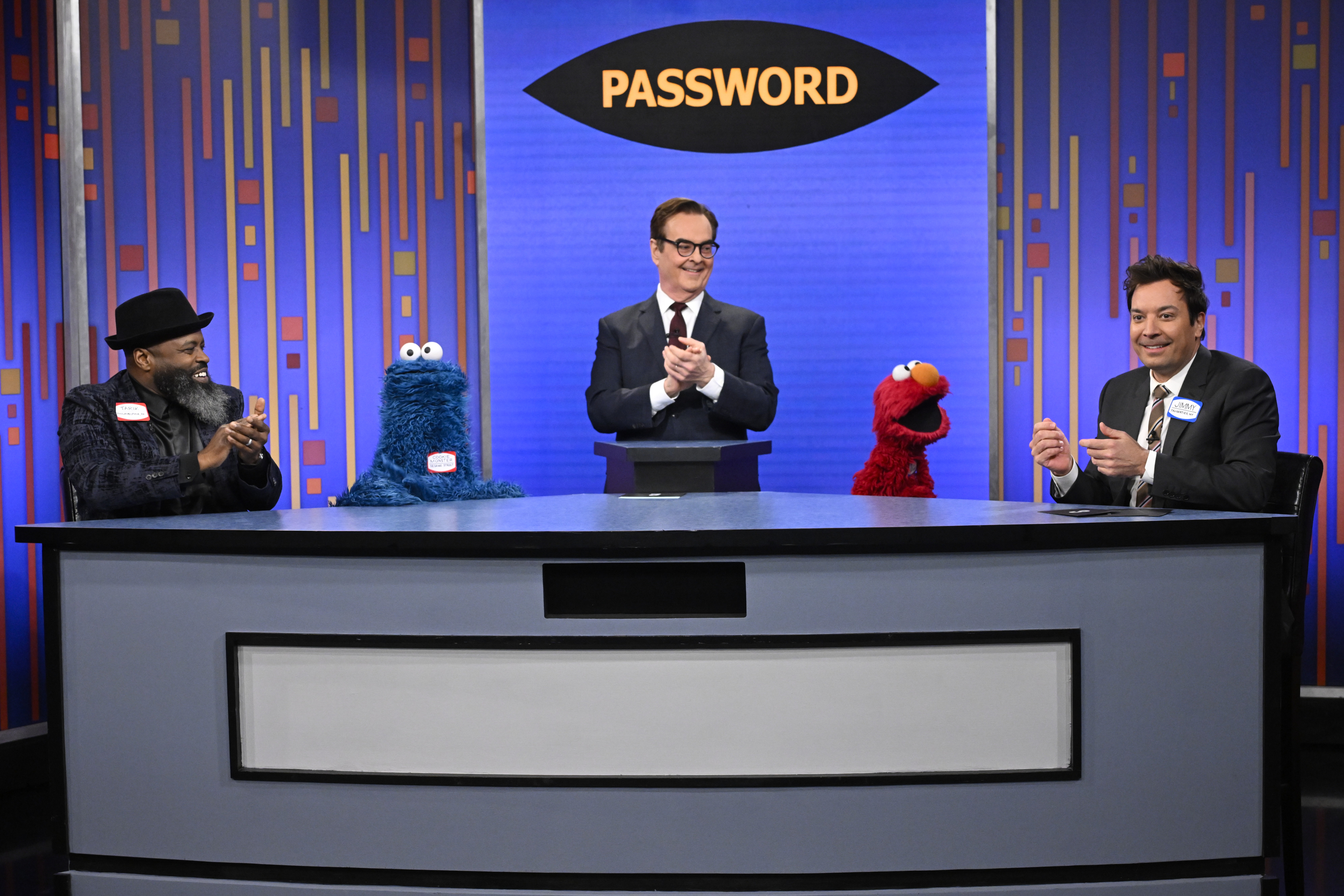Jimmy Fallon and guests, including Cookie Monster and Elmo, participate in a game show behind a desk with a sign that reads "Password."