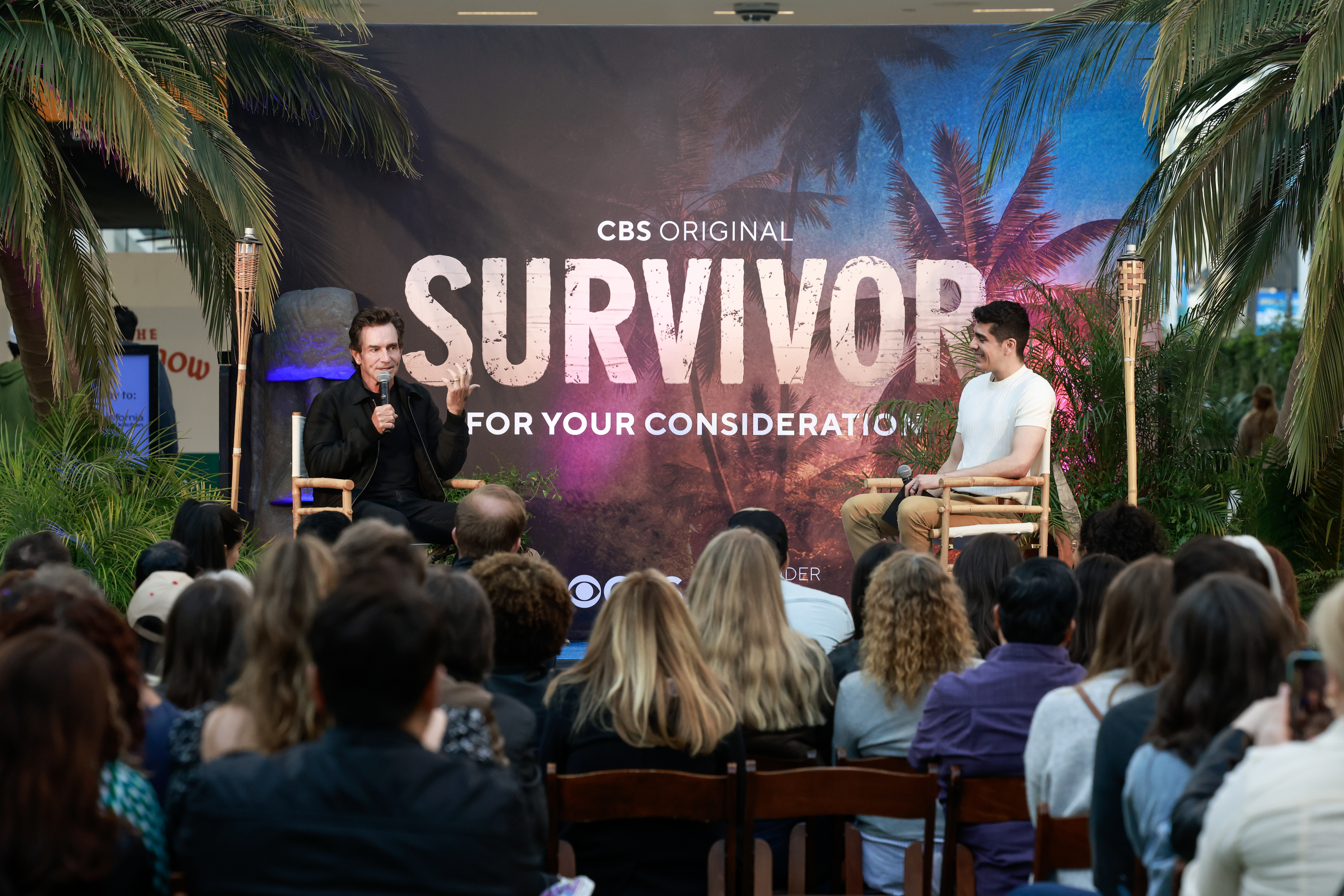 Two men sit on stage with a "Survivor" banner behind them, addressing an audience