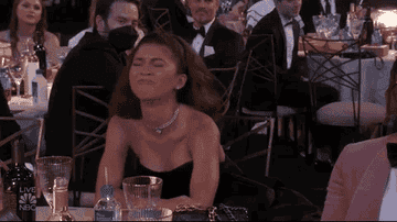 Zendaya sits at a table, smiling and laughing, wearing a strapless dress and a choker necklace at an event where others are also seated around tables