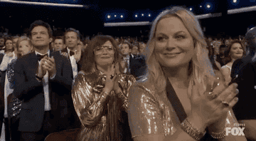 Amy Poehler and other audience members stand and clap at an awards show. Poehler smiles while wearing a shimmery dress