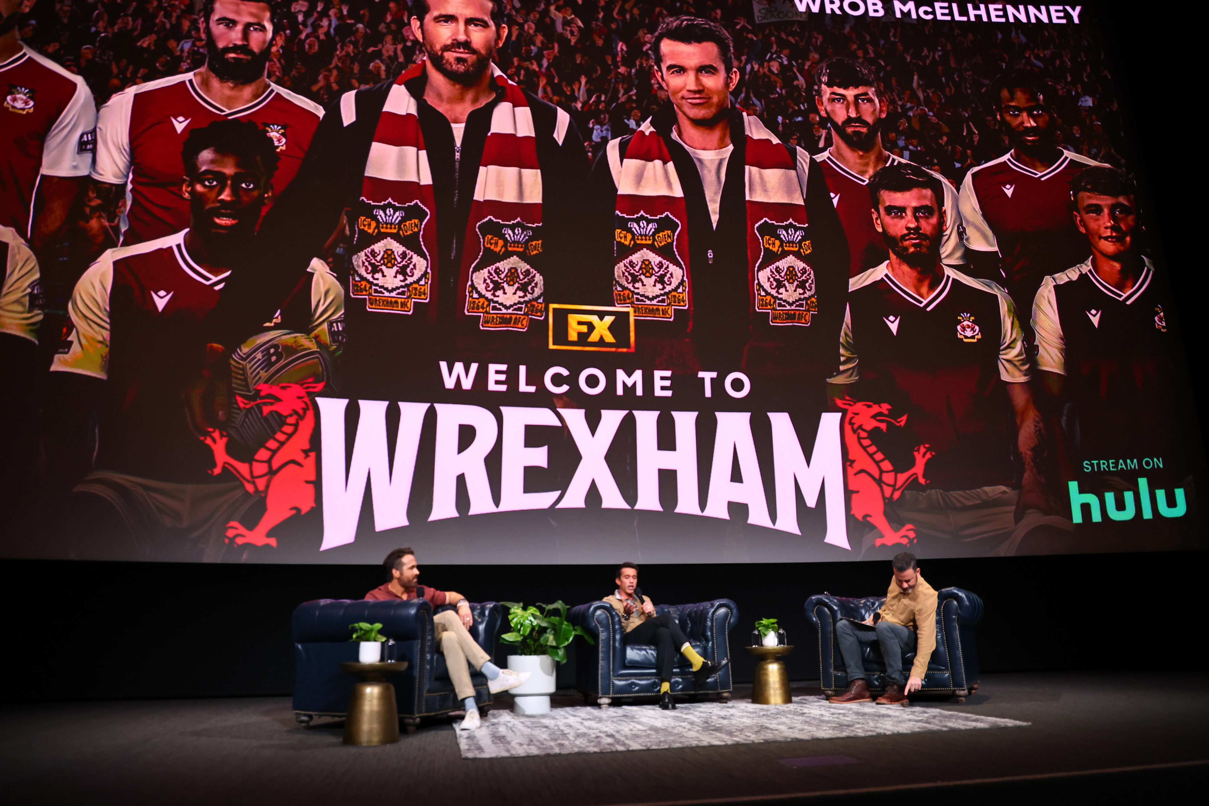 Three people seated on stage discuss "Welcome to Wrexham" in front of a large screen showing promotional material for the show