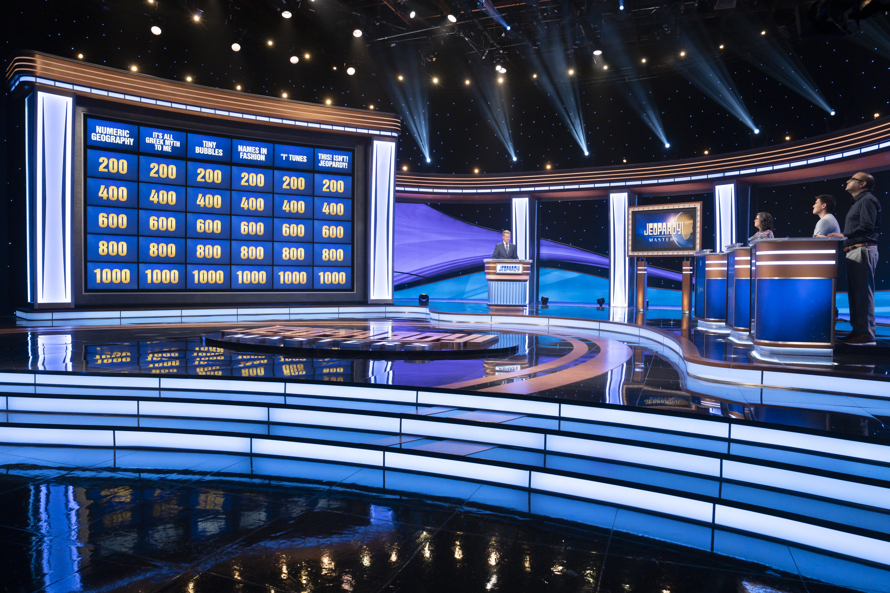 "Jeopardy!" game show set with three contestants and host. Contestants are at their podiums, and the game board displays categories and point values