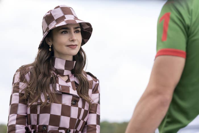 Lily Collins wears a checkered coat with a matching hat and gold earrings, talking to a person wearing a green shirt with a red number 1 on the sleeve