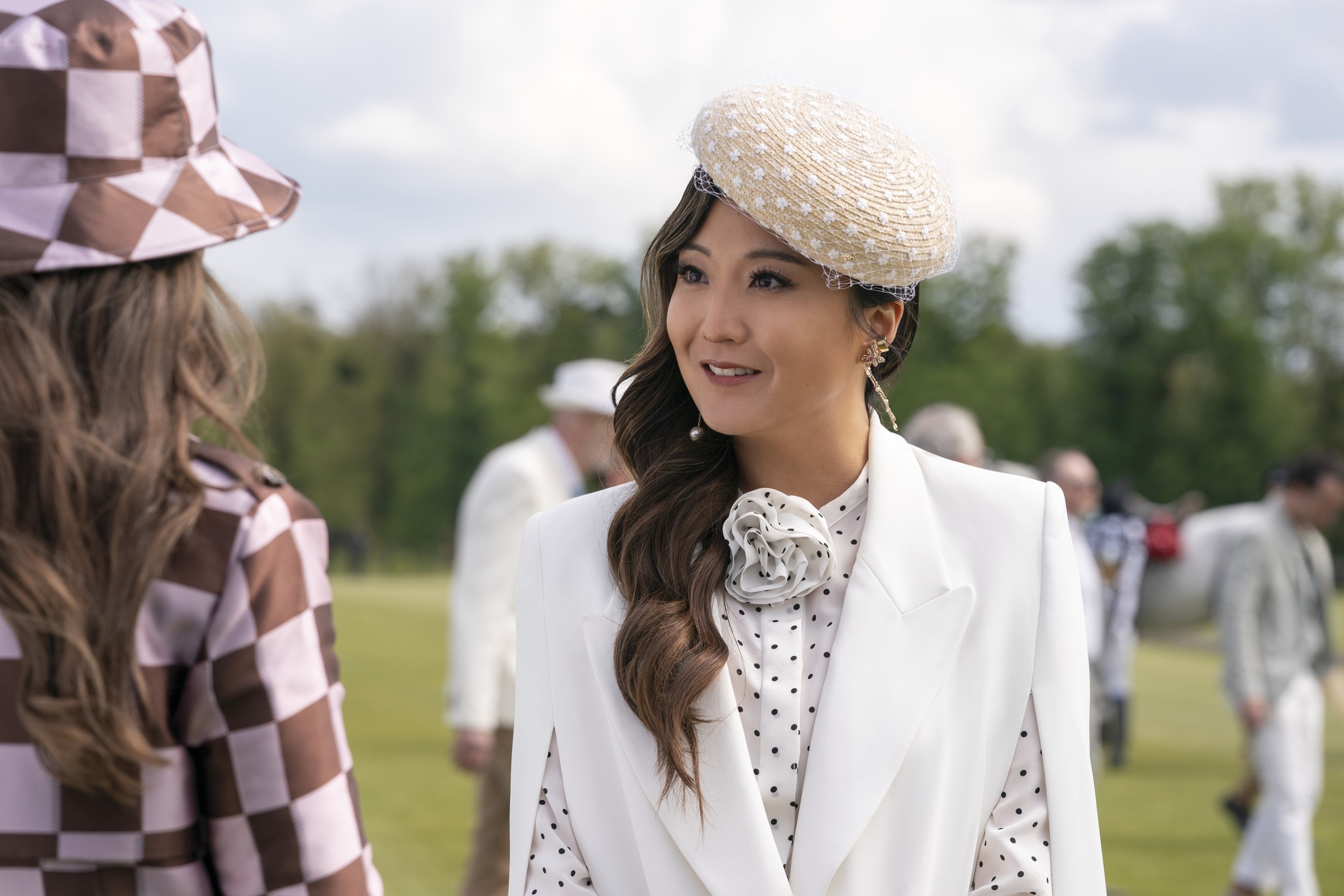 Ashley Park talking to another person at an outdoor event. She is wearing a white ensemble with a polka dot shirt, a large flower brooch, and a stylish hat