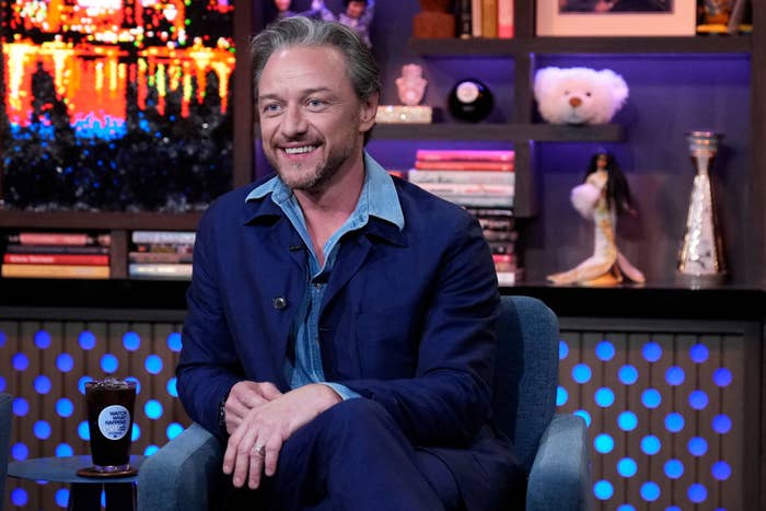 James McAvoy sits on a talk show set, smiling, wearing a blue jacket and shirt. Shelves with books and decor are visible in the background