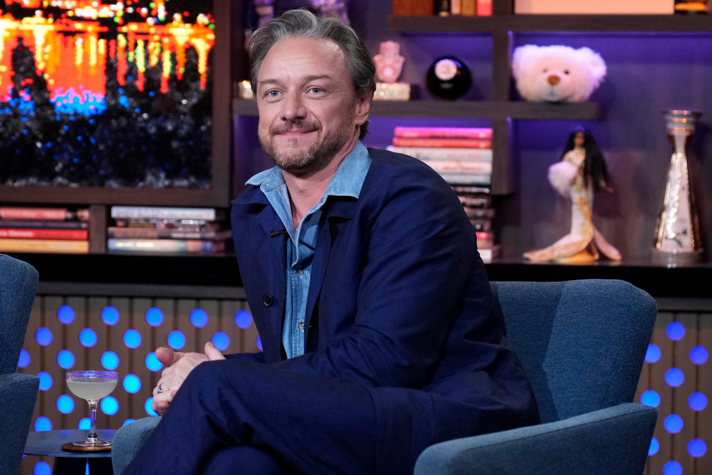 James McAvoy sits on a talk show set, sporting a blue jacket over a light shirt, with a relaxed and smiling demeanor