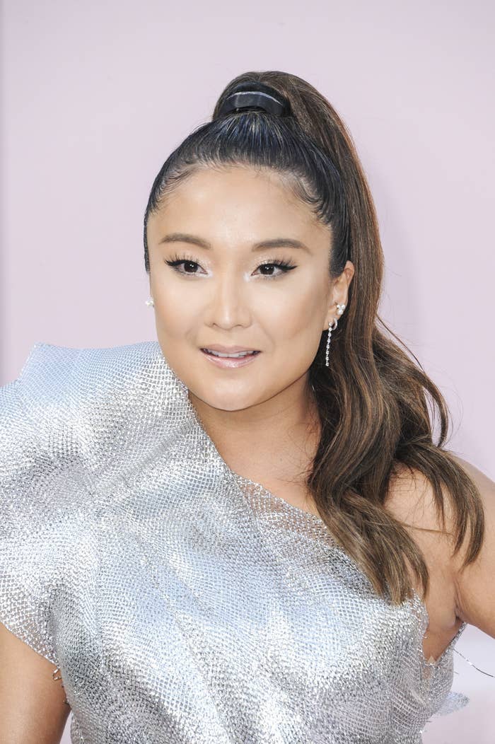 Woman with long hair in a high ponytail wears a one-shoulder, silver textured dress, smiling at the camera