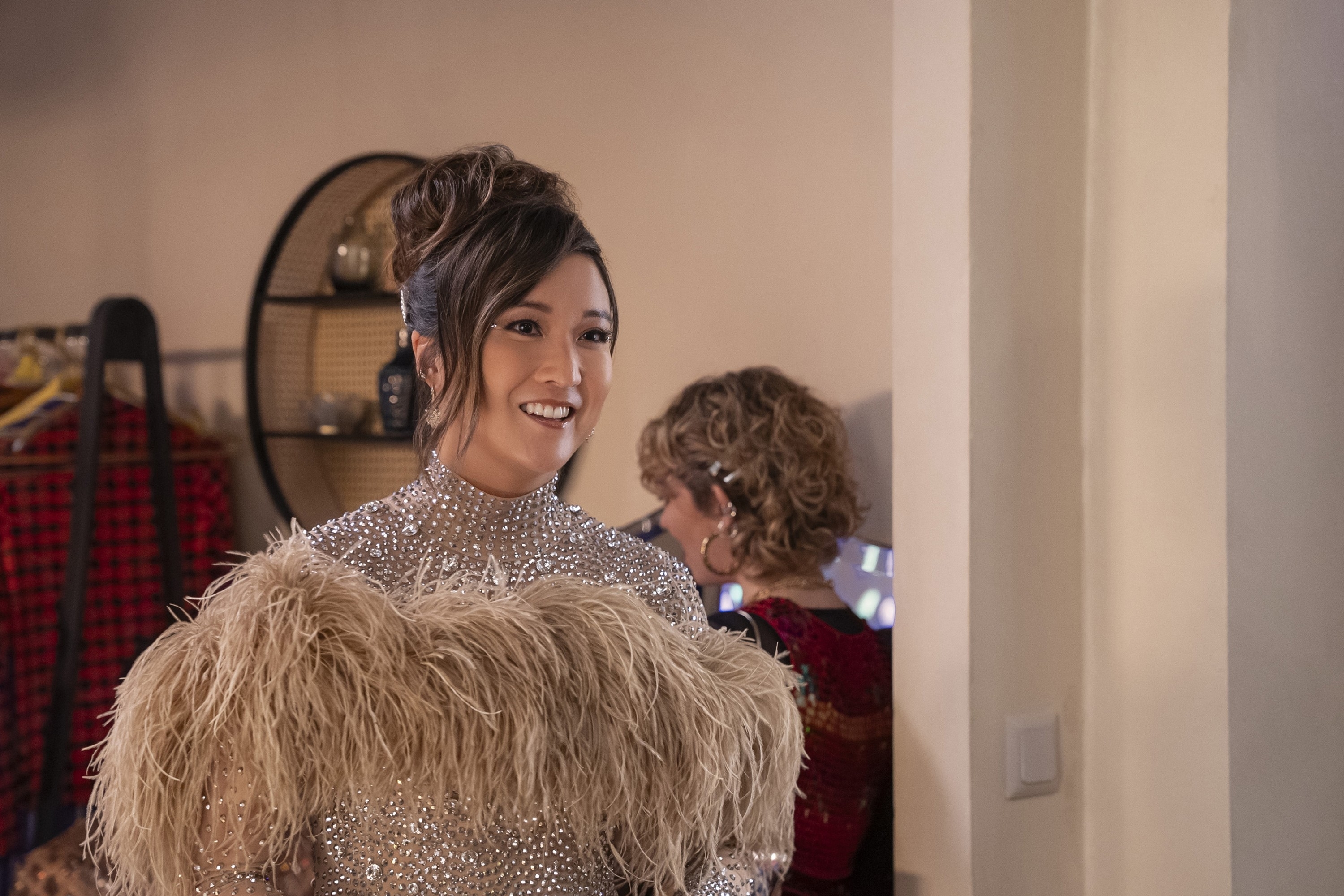 Ashley Park in a sparkly dress with a feathered accessory, smiling and looking to the side. Another person in the background is partially visible