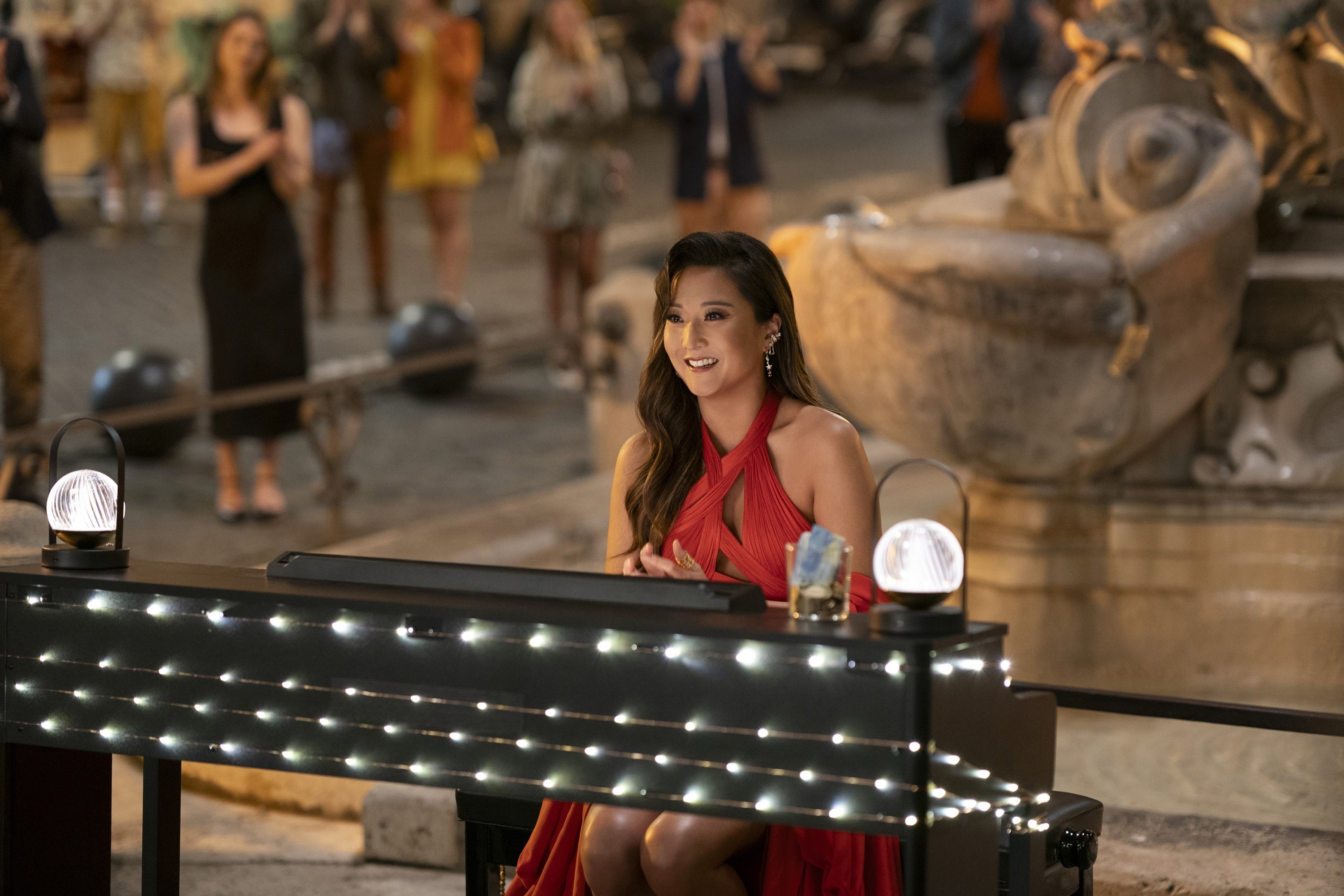 Woman in a red dress playing a piano in an outdoor setting, with onlookers in the background