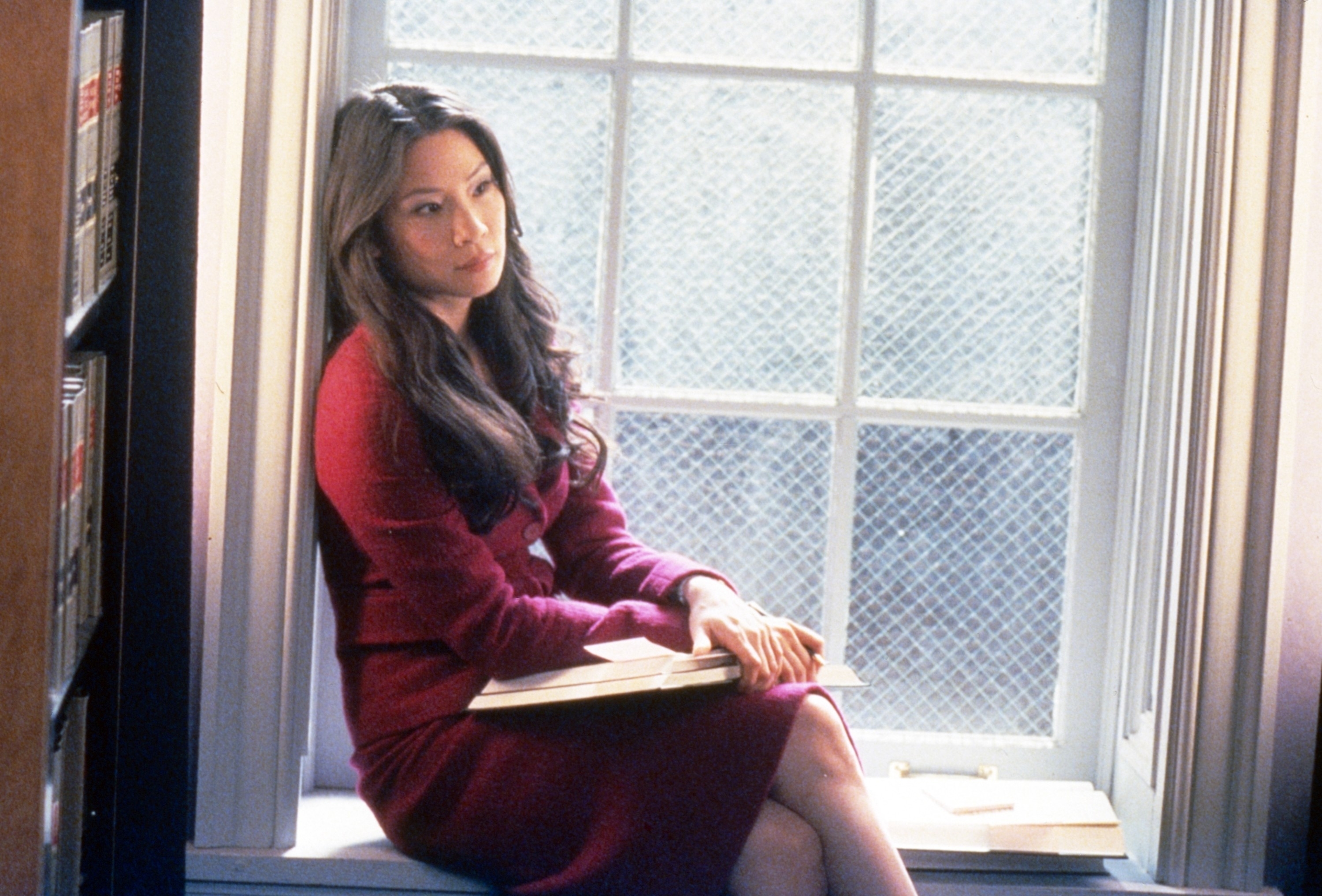 Lucy Liu sits by a window, holding a book in her lap. She wears a stylish, knee-length dress