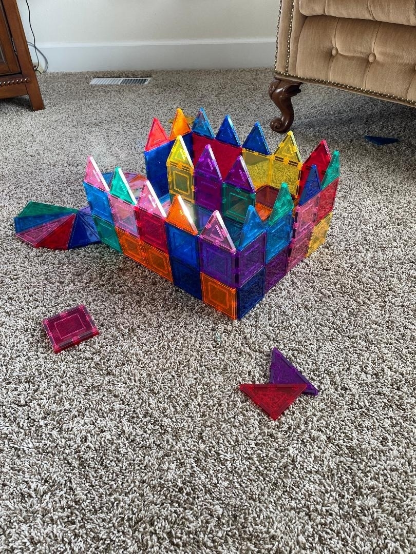 A castle-like structure made of colorful magnetic tiles is built on a carpet. A few tiles are scattered around it. No people are present