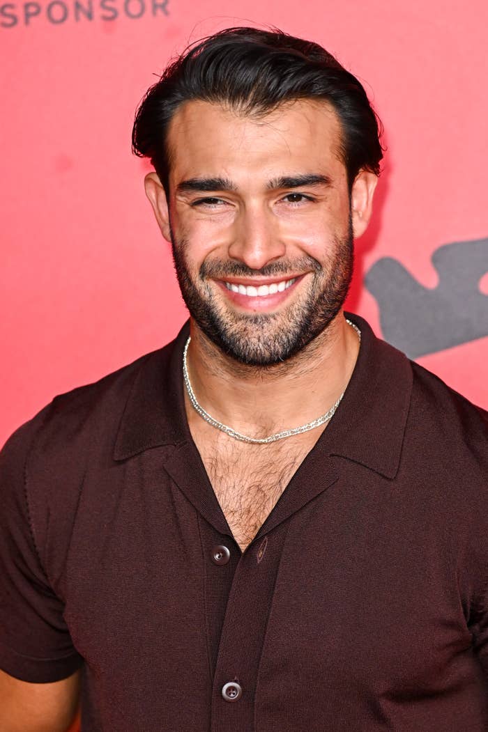 Man smiling on a red carpet event, wearing a short-sleeved shirt and chain necklace