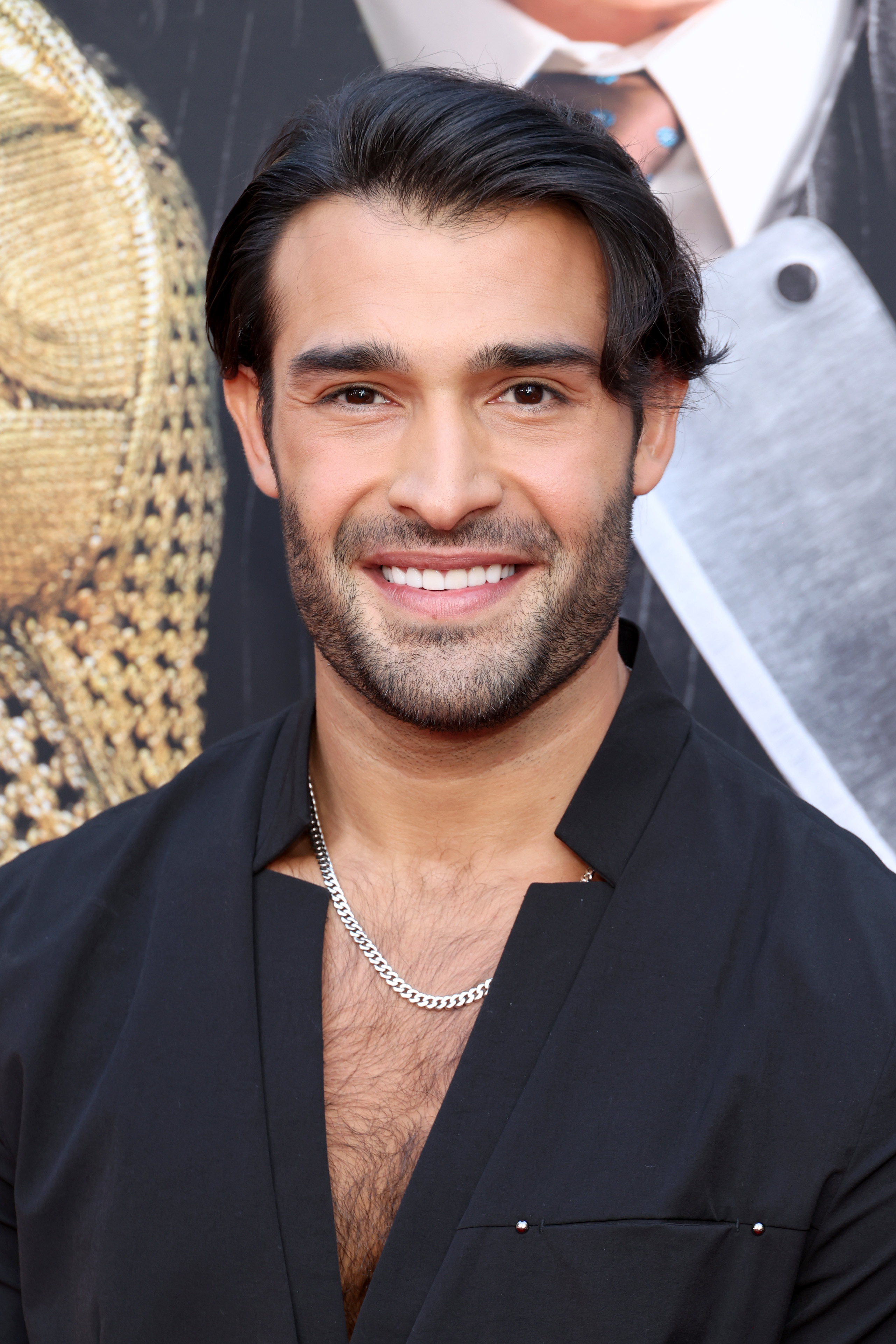 Man smiling in front of a backdrop, wearing a black open-neck shirt and a silver chain necklace
