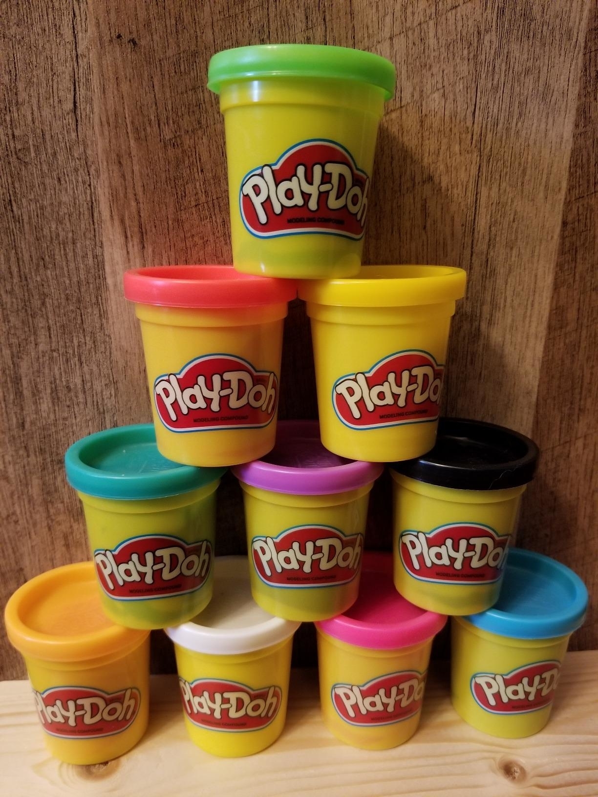 Nine Play-Doh containers in various colors, stacked in a pyramid on a wooden surface, used in an article about shopping for children’s toys
