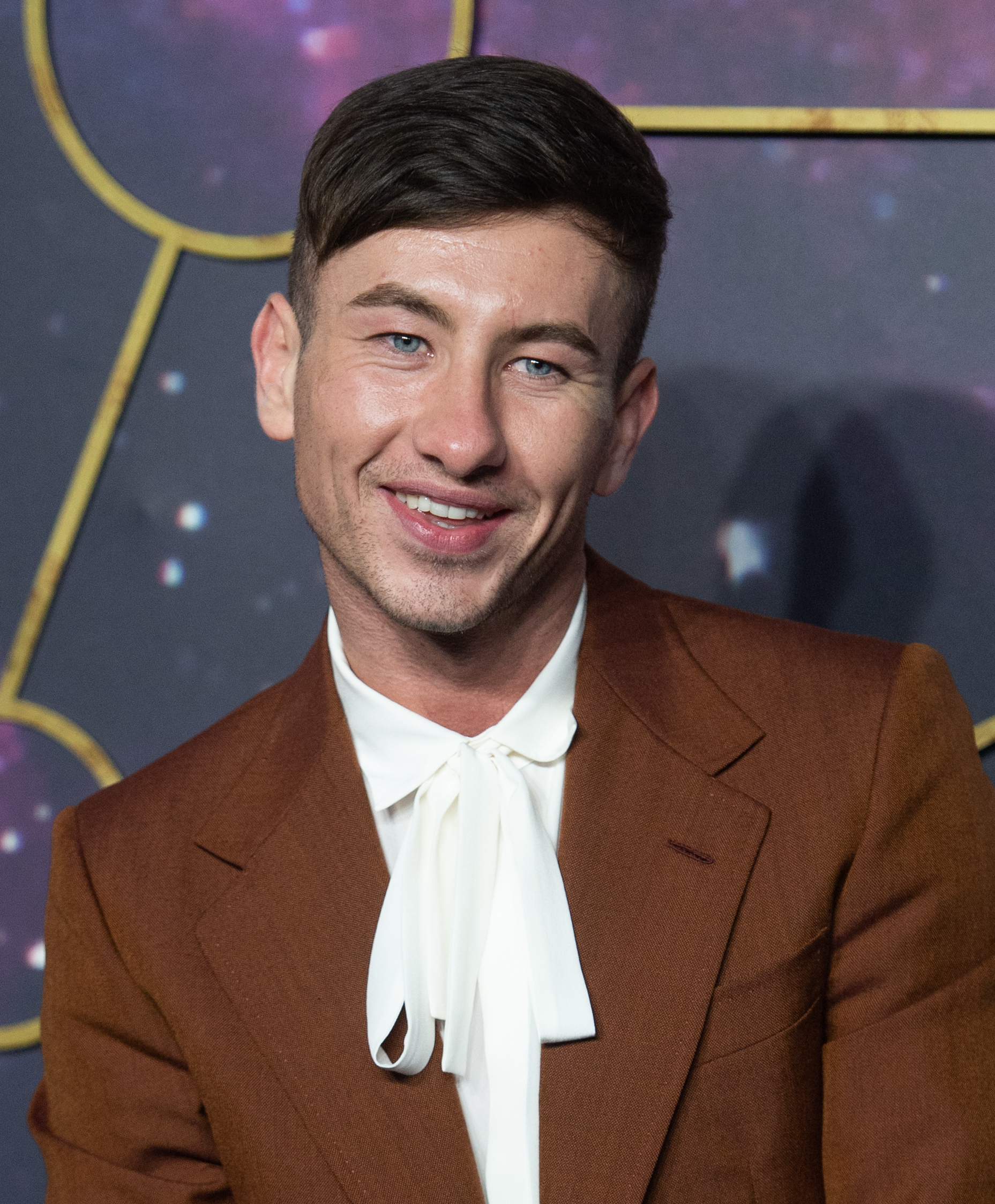 Barry Keoghan smiling, wearing a brown suit and white bow-tie, posing at a formal event