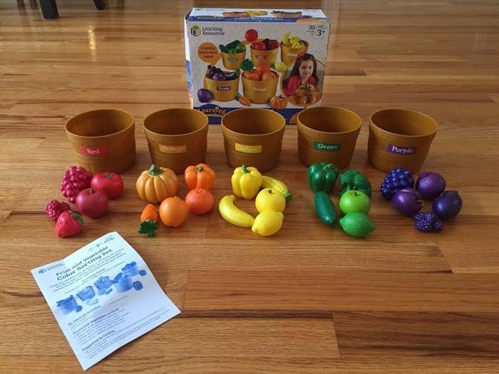 Toy set with five brown bowls labeled by colors, various plastic fruits and vegetables, a leaflet, and packaging featuring a young girl playing with the set
