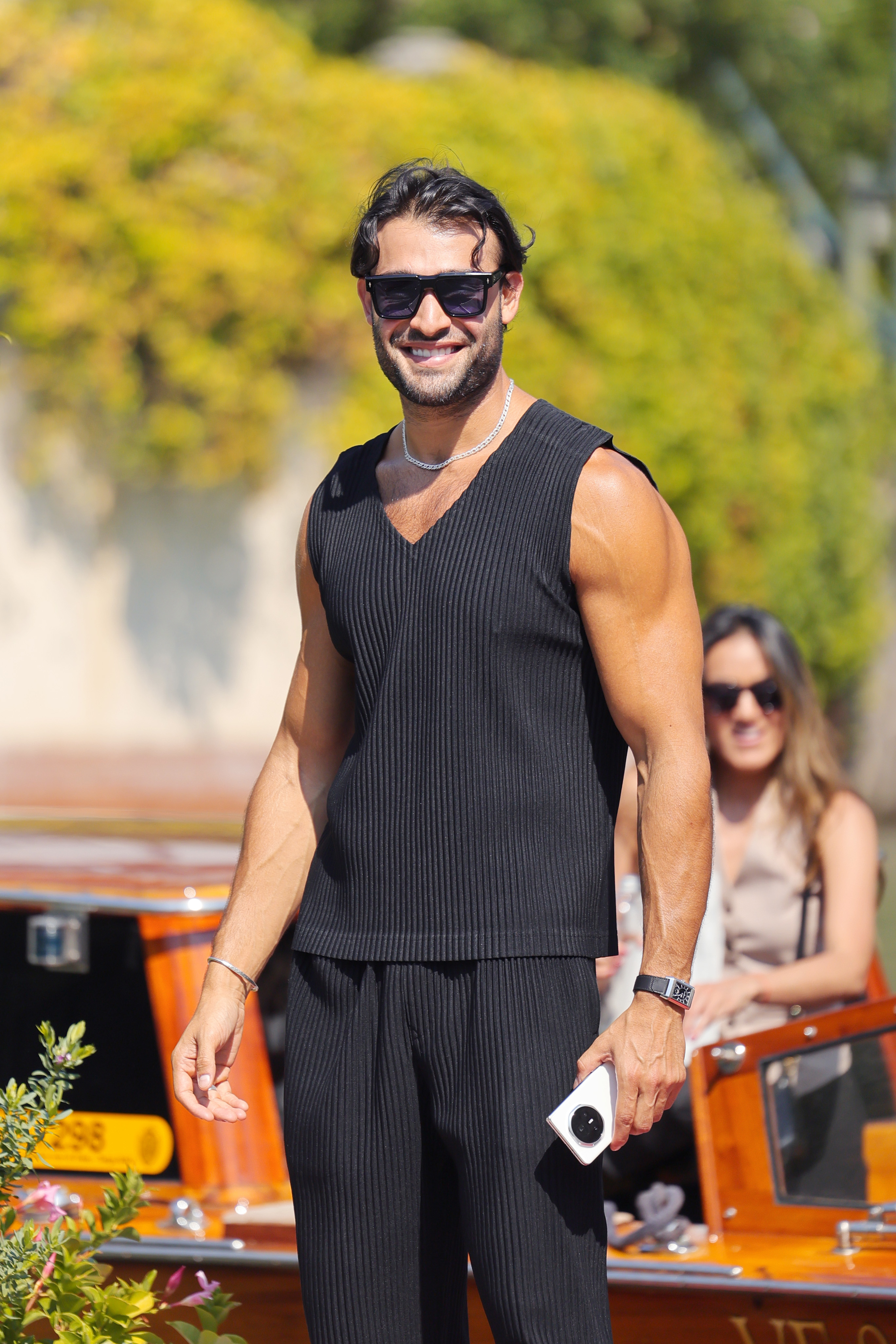 Man in sleeveless, V-neck top and pants, wearing sunglasses and holding a camera. Woman in the background on a boat. Names unknown